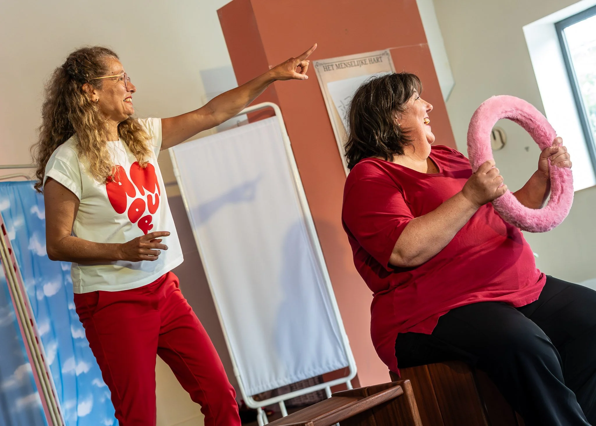 Twee vrouwen, een in een witte T-shirt met rode tekst en een in een rode blouse, lachen en maken een gebaar tijdens een activiteit op een kamer met raam en muurdecoraties.
