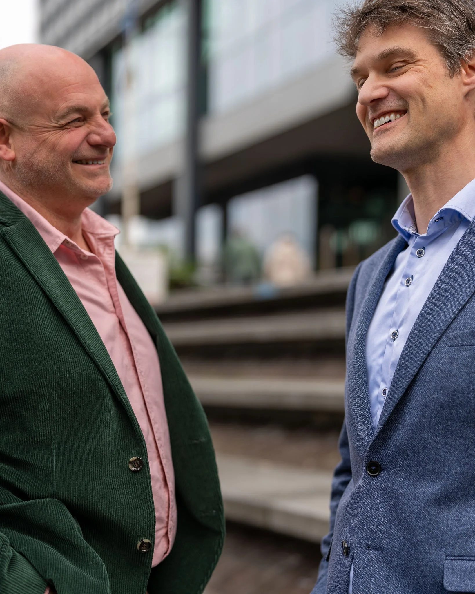 Twee mannen glimlachen tijdens een gesprek, ze dragen nette blazers, de ene in groen en de andere in blauw, buiten voor een modern gebouw.