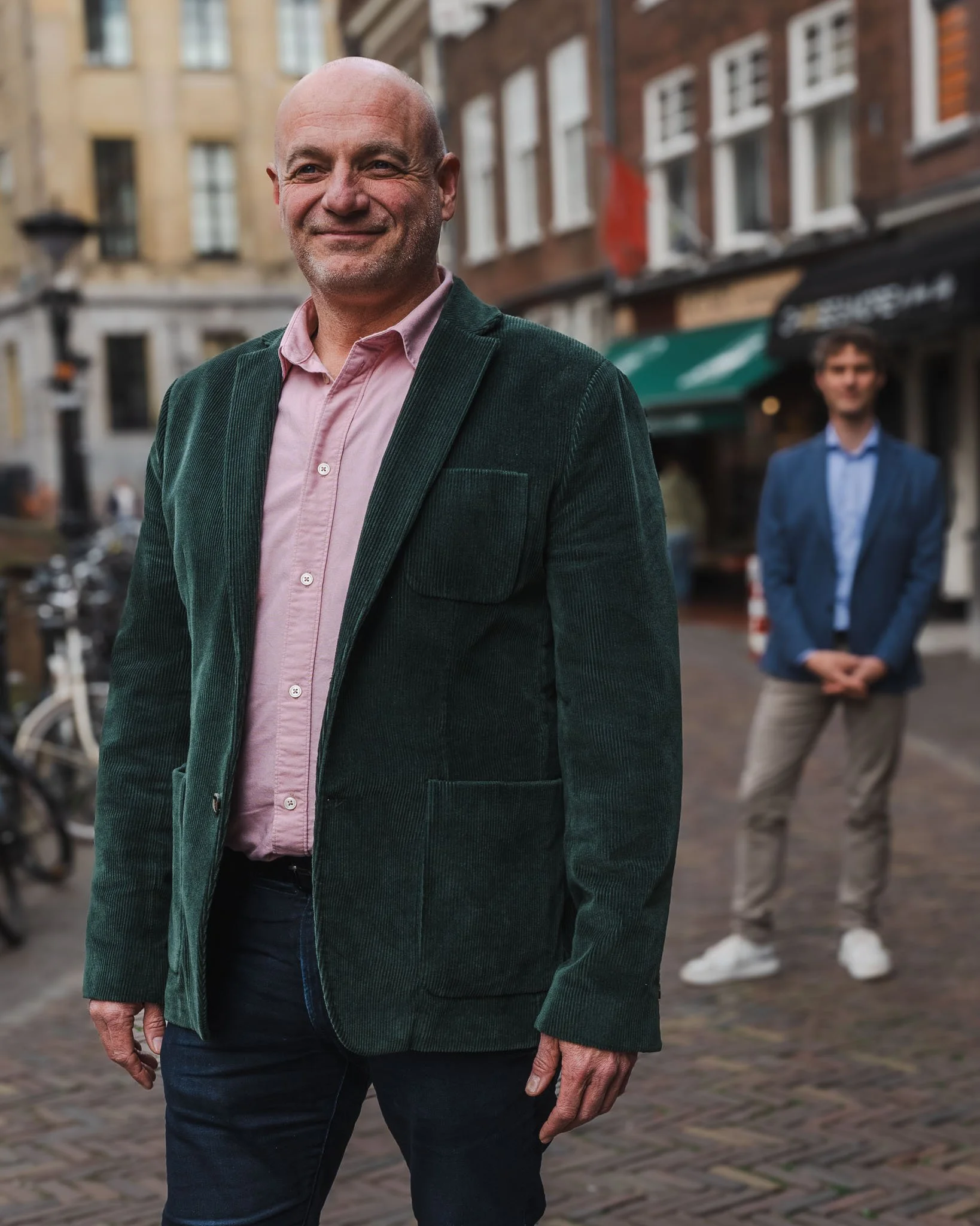 Een oudere man in een groen jasje en roze overhemd die glimlacht op een straat met stenen pavement, met een andere man op de achtergrond.