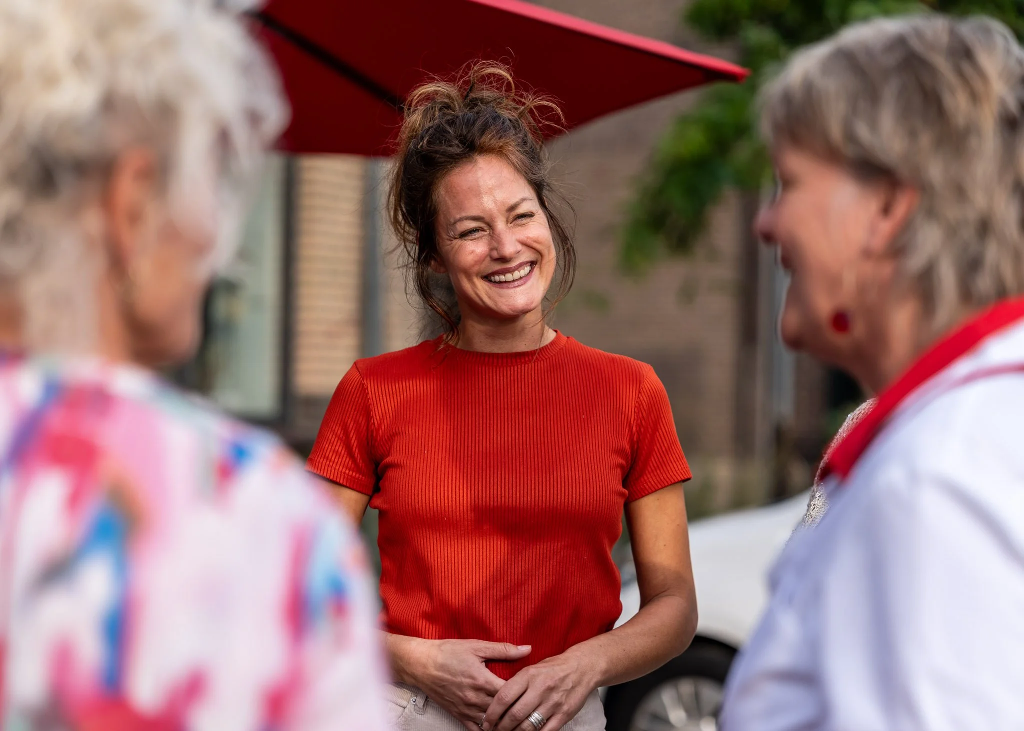 Vrouw in rood shirt glimlachend tijdens gesprek met groep ouderen buiten onder een parasol.