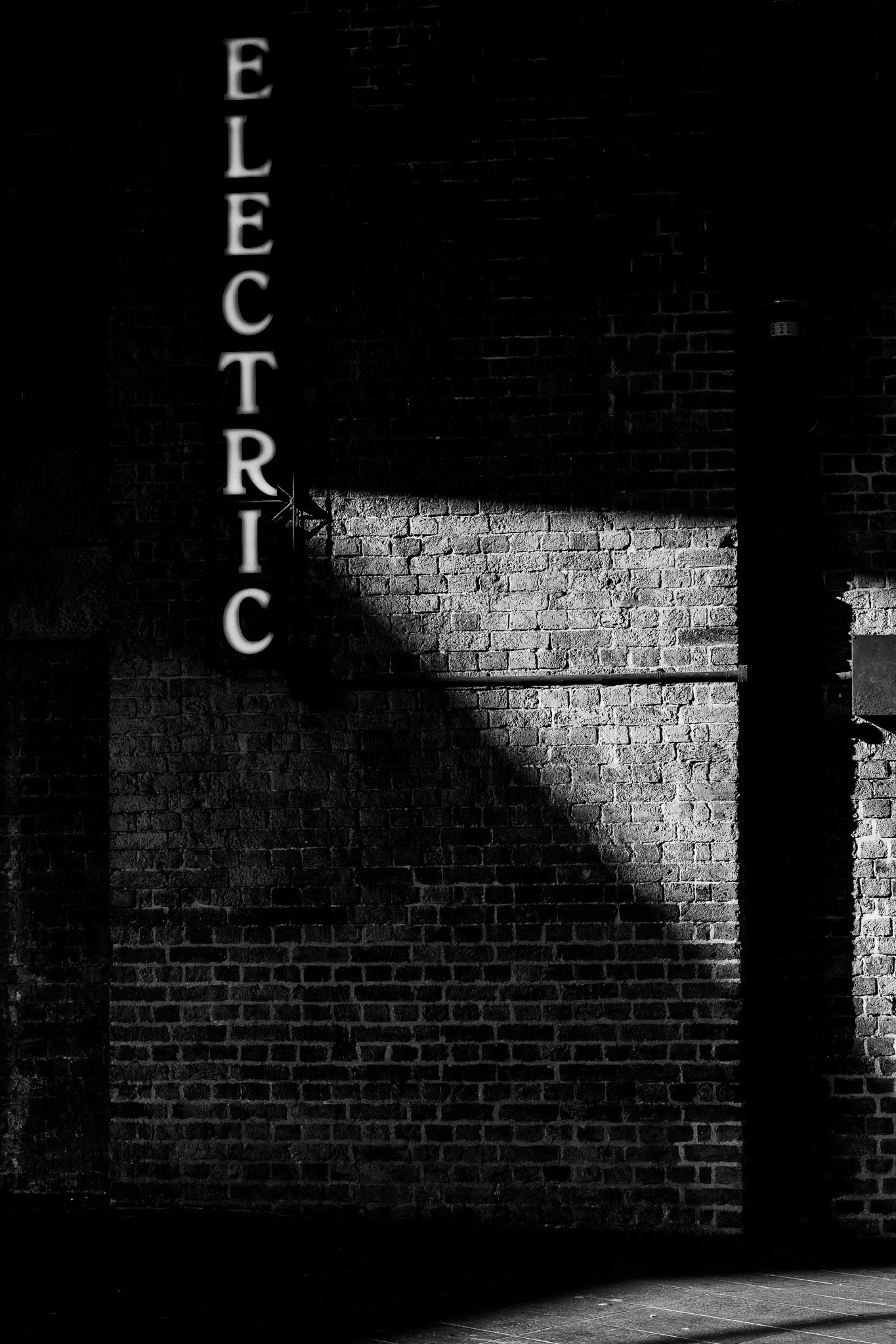Een zwart-wit foto van een bakstenen muur met een verlichte neonbalk met de tekst 'ELECTRIC' en richtingaanwijzer.
