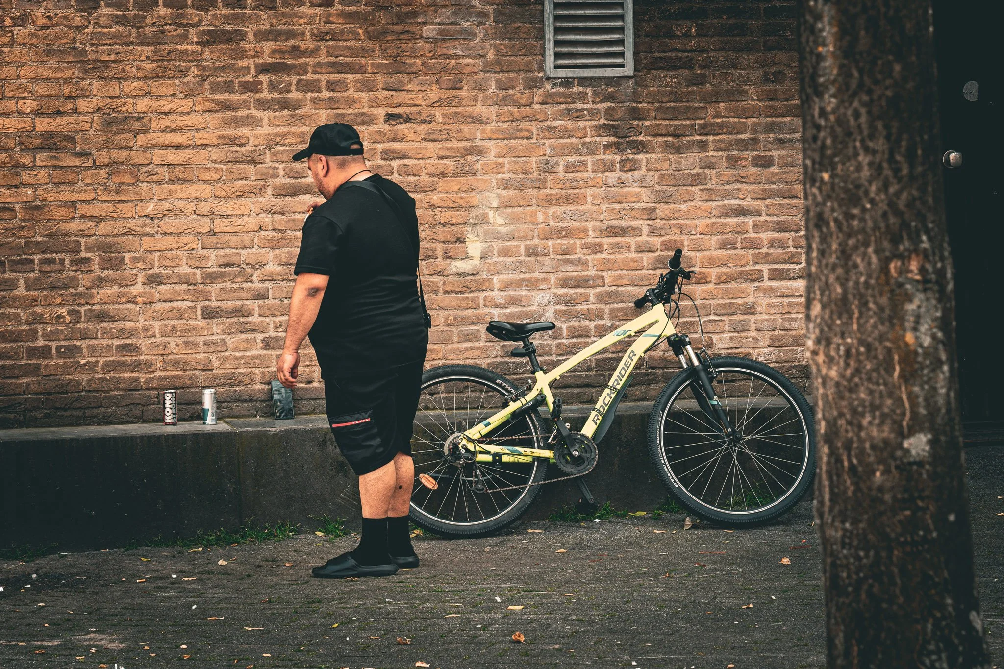 Man staat naast een gele mountainbike tegen een bakstenen muur, met een koeler, een blikje en een flesje op de rand.
