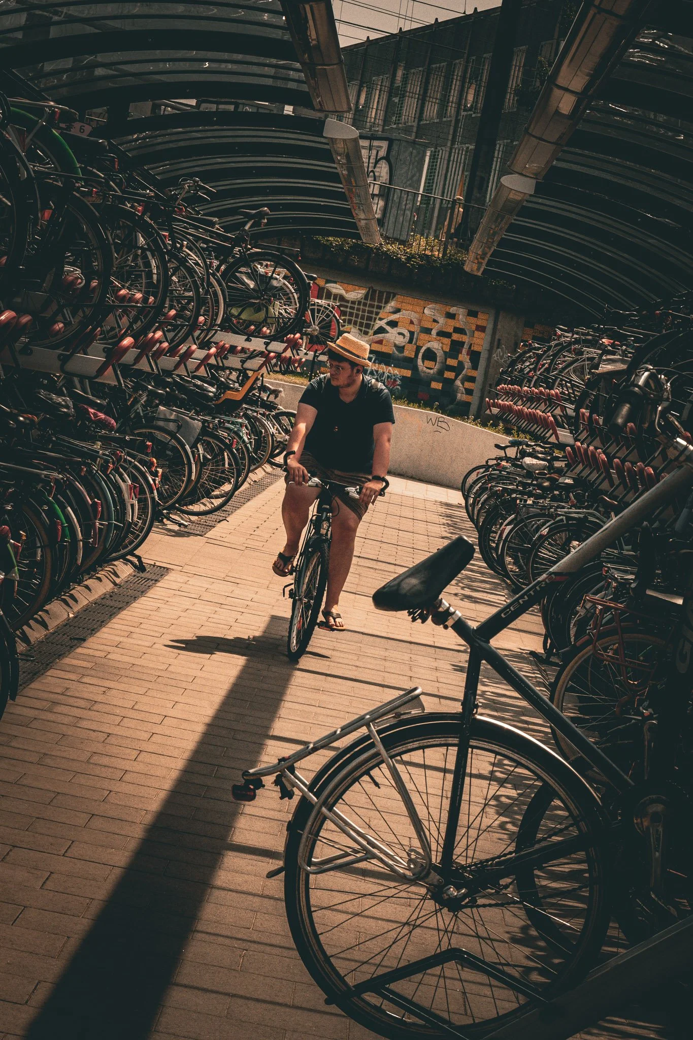 Man fietst door een fietsenstalling met veel gestapelde fietsen, onder een overdekte brug met graffiti op de muur en moderne architectuur.