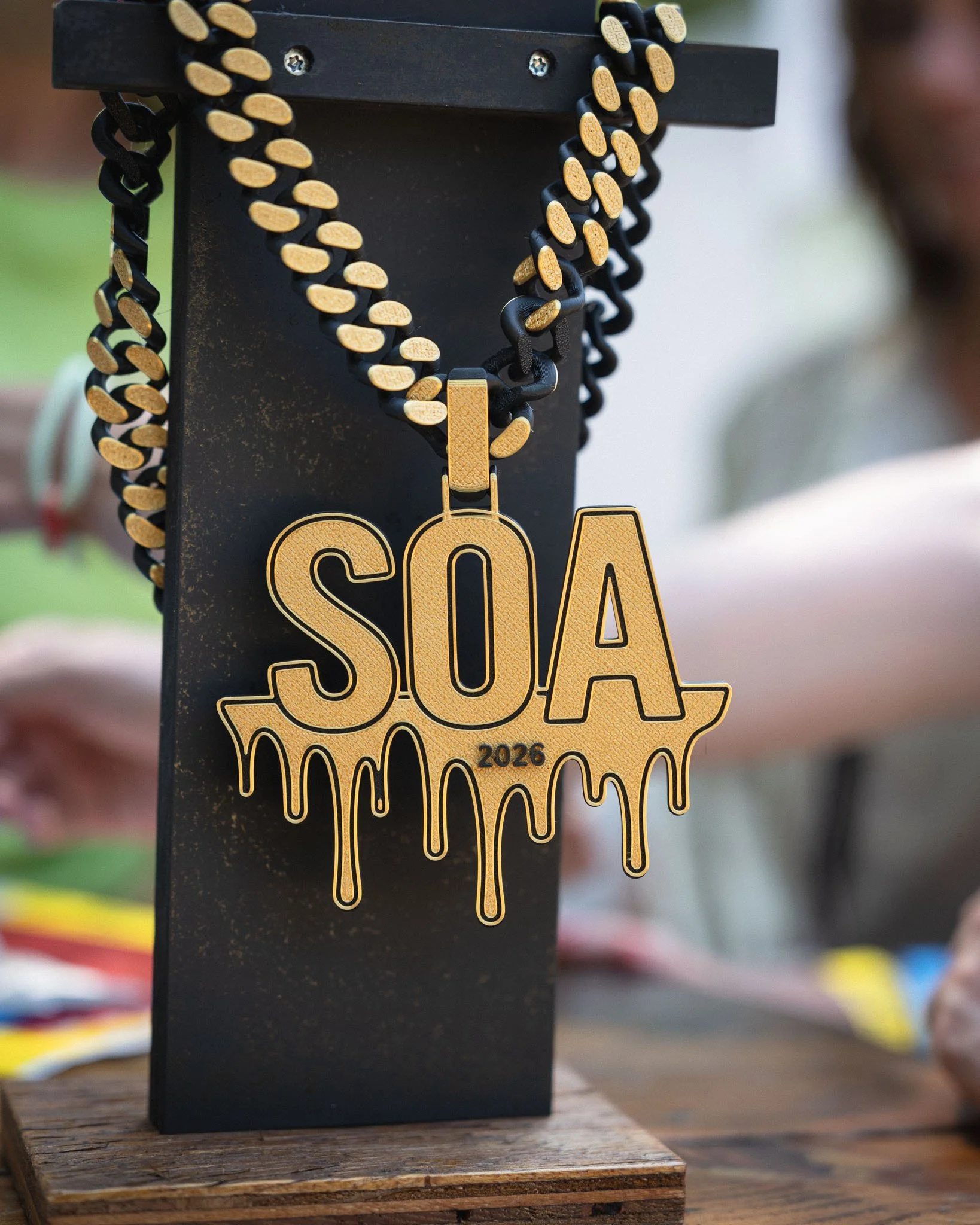 Een versierd medaille met de tekst 'SOTA 2026' en een goudkleurig ontwerp dat eruitziet als gesmolten goud, hangt aan een zwarte houten standaard.