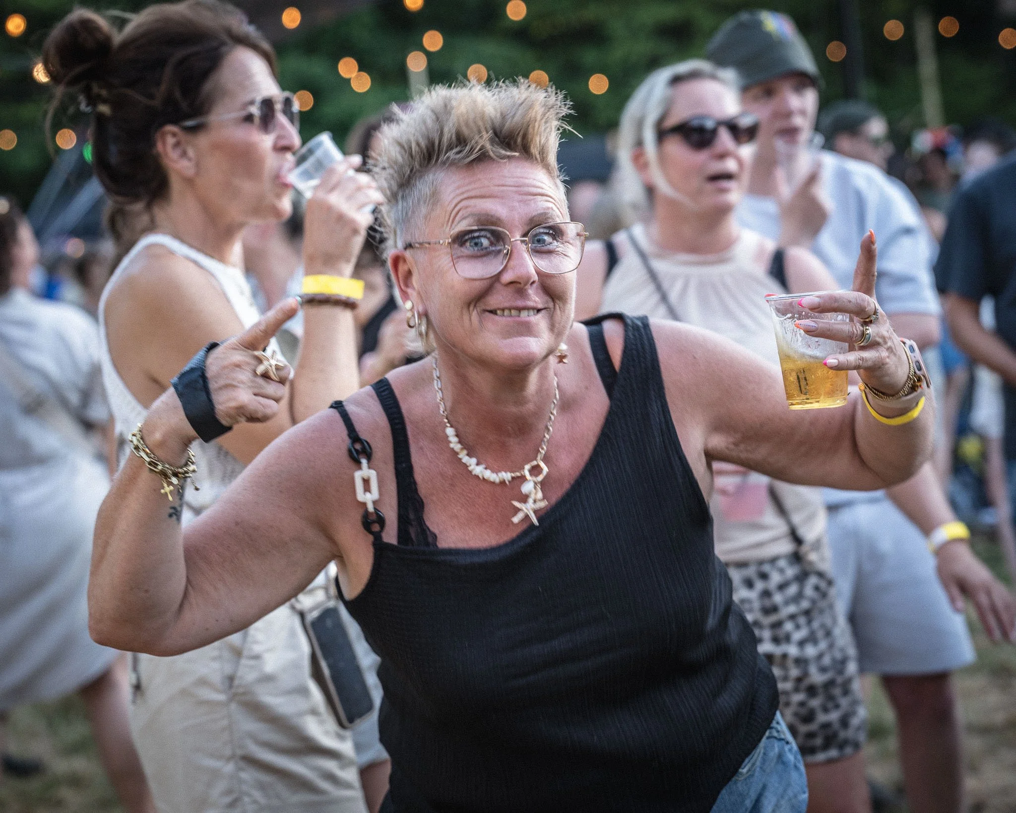 Groep mensen die op een festival of buitenfeest samen genieten, met één vrouw in de voorgrond die een drankje vasthoudt en glimlacht.
