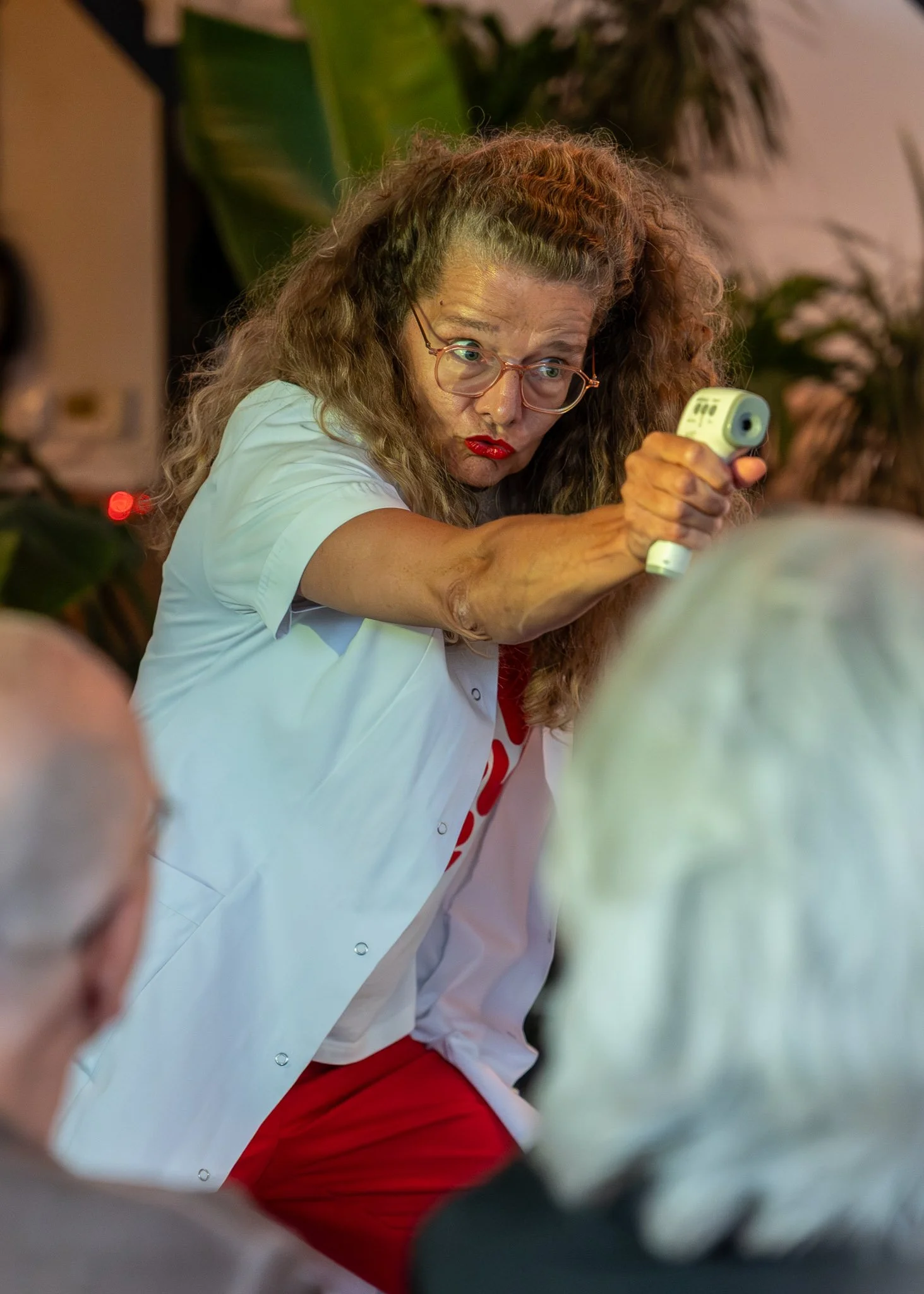 Een vrouw met krullend haar en een bril, gekleed in een wit en rood uniform, die haar hand uitstrekt met een medisch apparaat, waarschijnlijk een bloeddrukmeter, in een kamerverpakking met planten op de achtergrond.