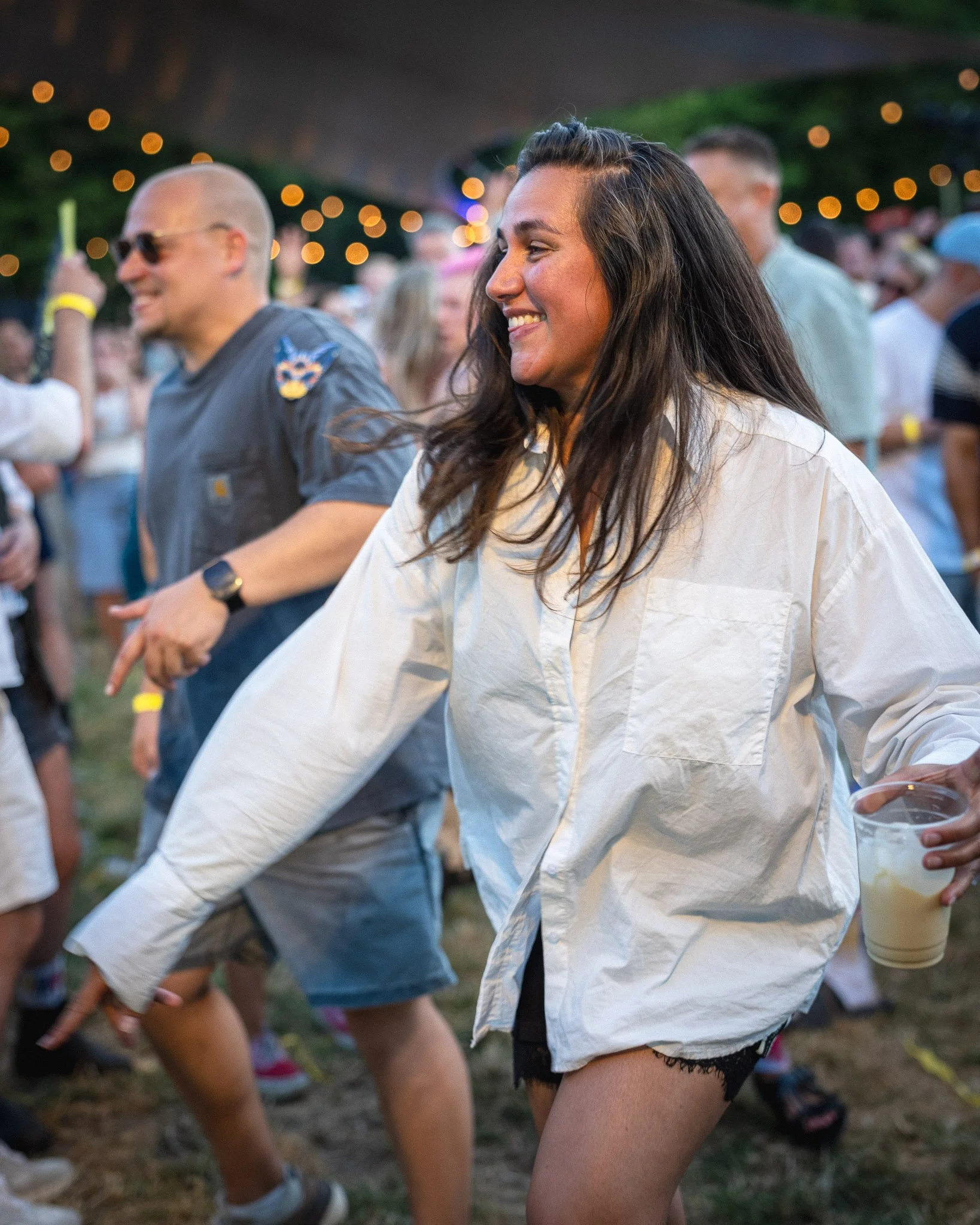 Mensen dansen op een festival in de avond, glimlachend en plezier hebbend, met feestverlichting op de achtergrond.