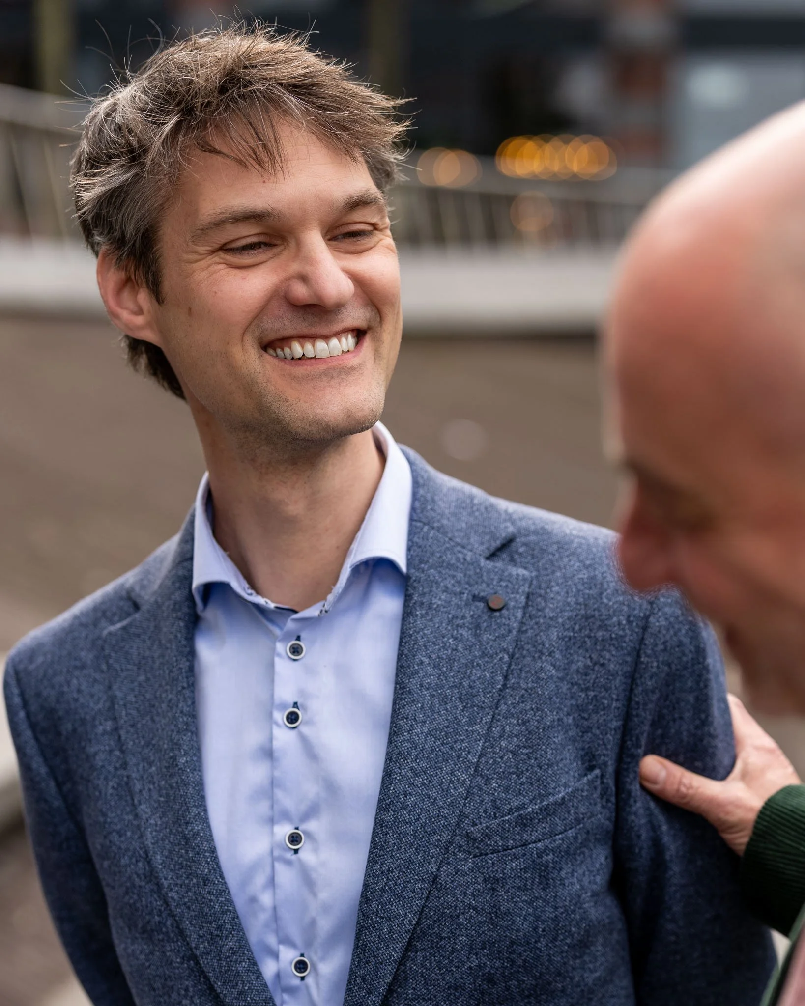Lachende man in een grijsblauw pak wordt begroet door een andere persoon, die een hand op zijn schouder legt, op een outdoorterras met stedelijke achtergrond.
