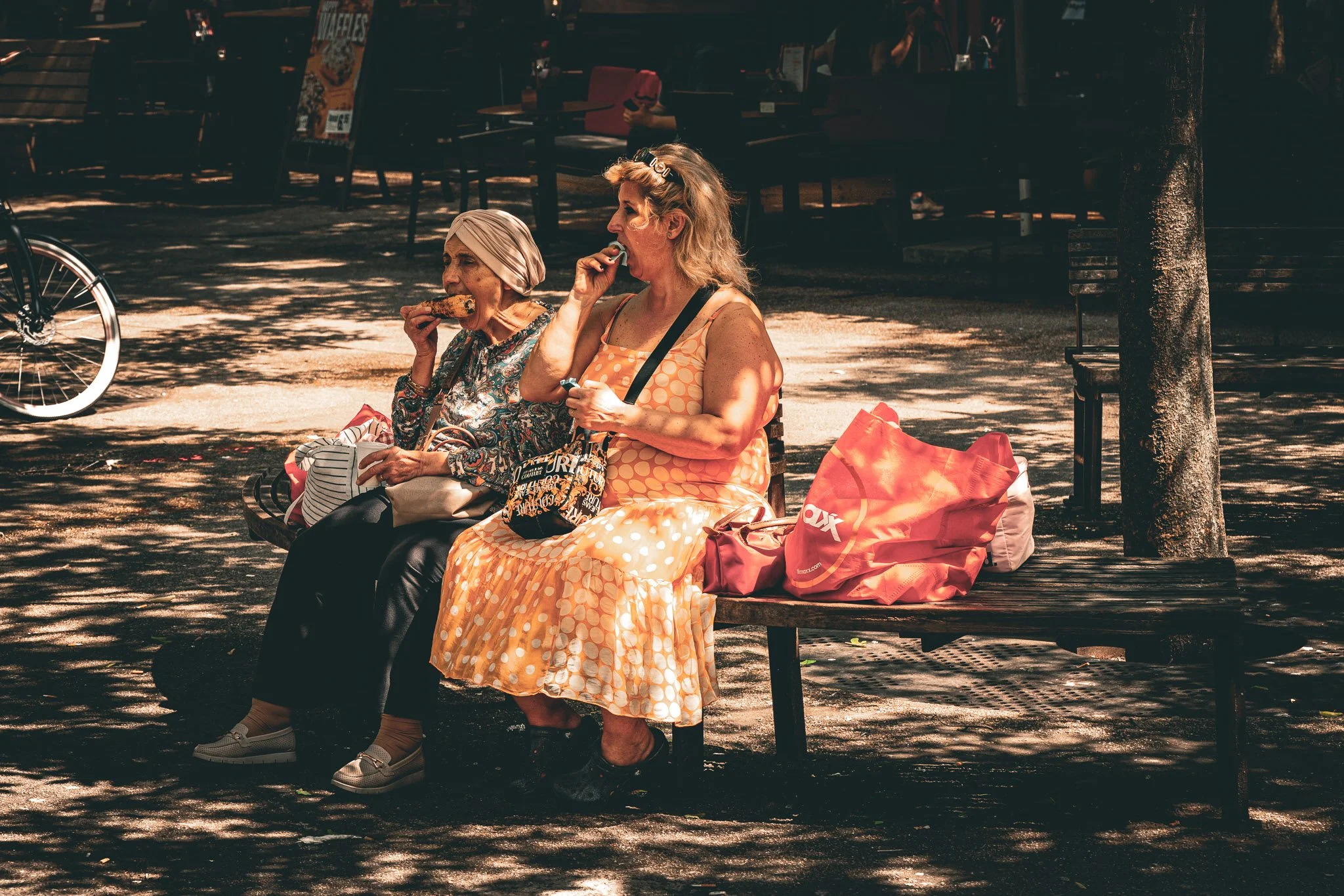 Twee vrouwen zitten op een bank in een park, eten wat snacks in de schemering met schaduw van bladeren boven hen.