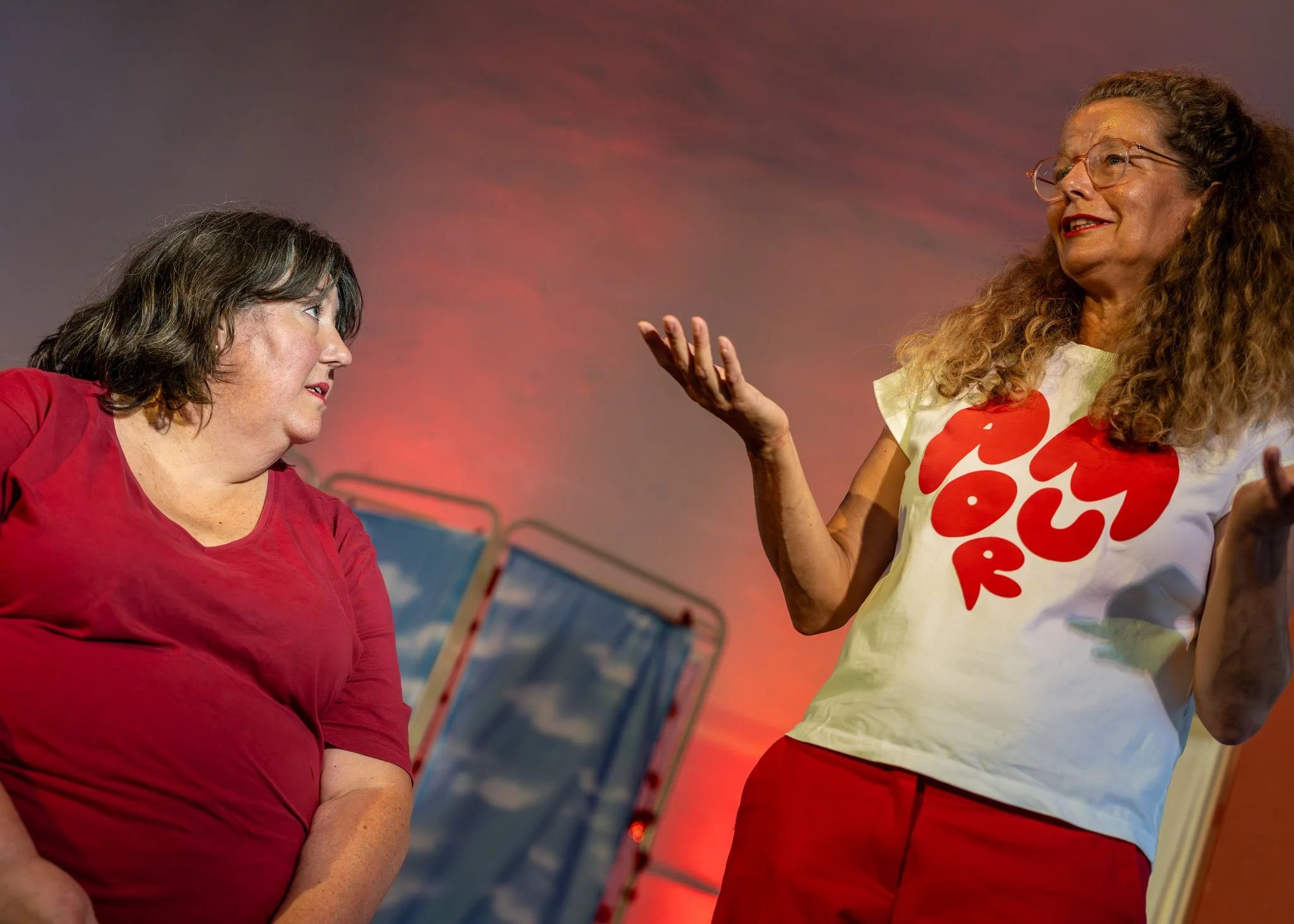 Twee vrouwen in gesprek, één kijkt serieus en de andere glimlacht, binnen een ruimte met kleurrijke achtergrond en banners.