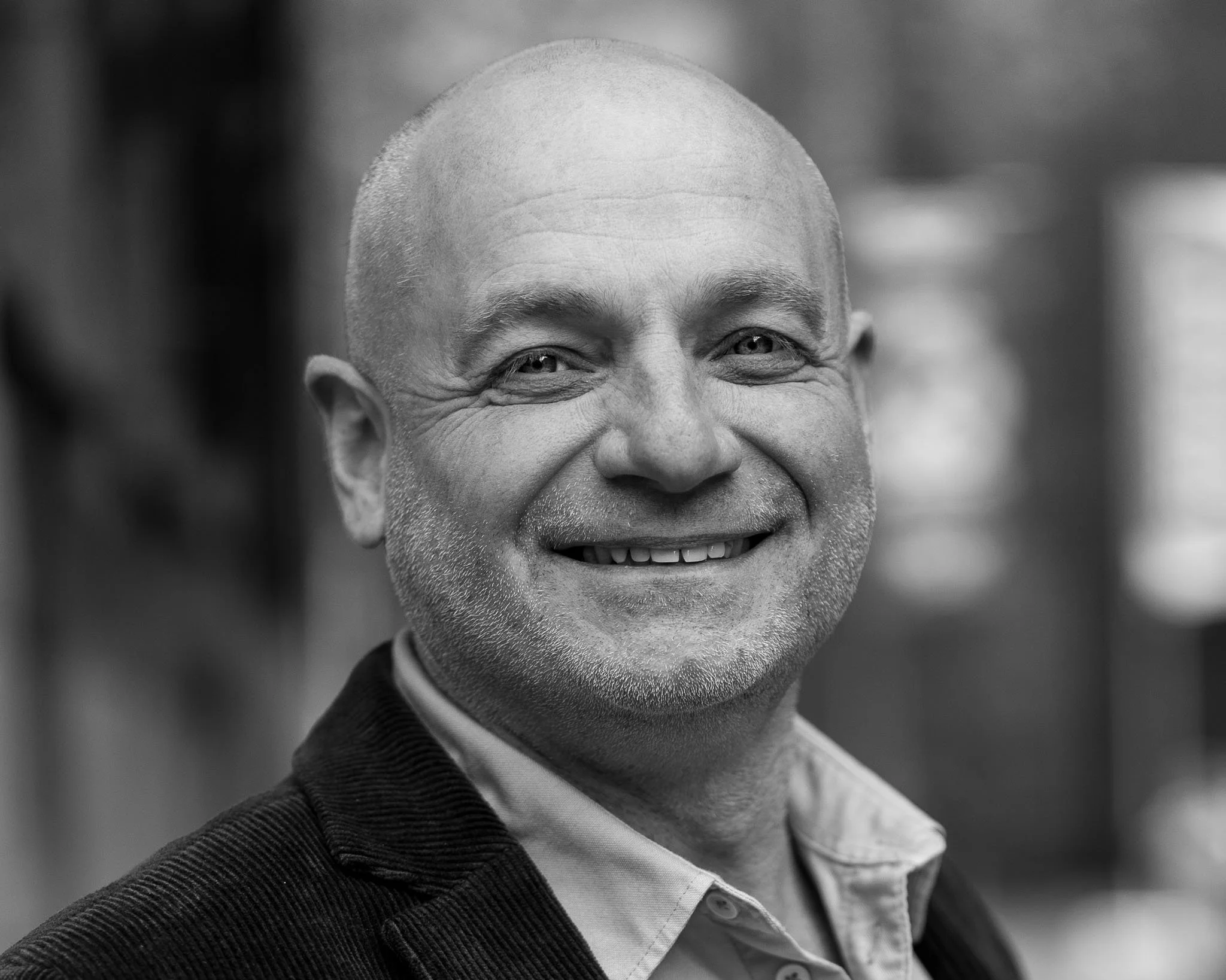 Portret van een glimlachende man met een kale kop, gekleed in een donker colbert en een witte blouse, op een vage achtergrond.