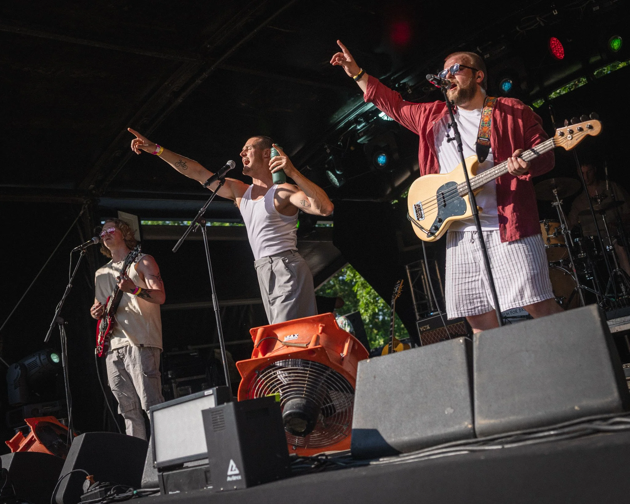 Muzikanten op een podium tijdens een live optreden, met één man die gitaar speelt en een andere man die zingt en de microfoon vasthoudt, onder kleurrijke lichten.