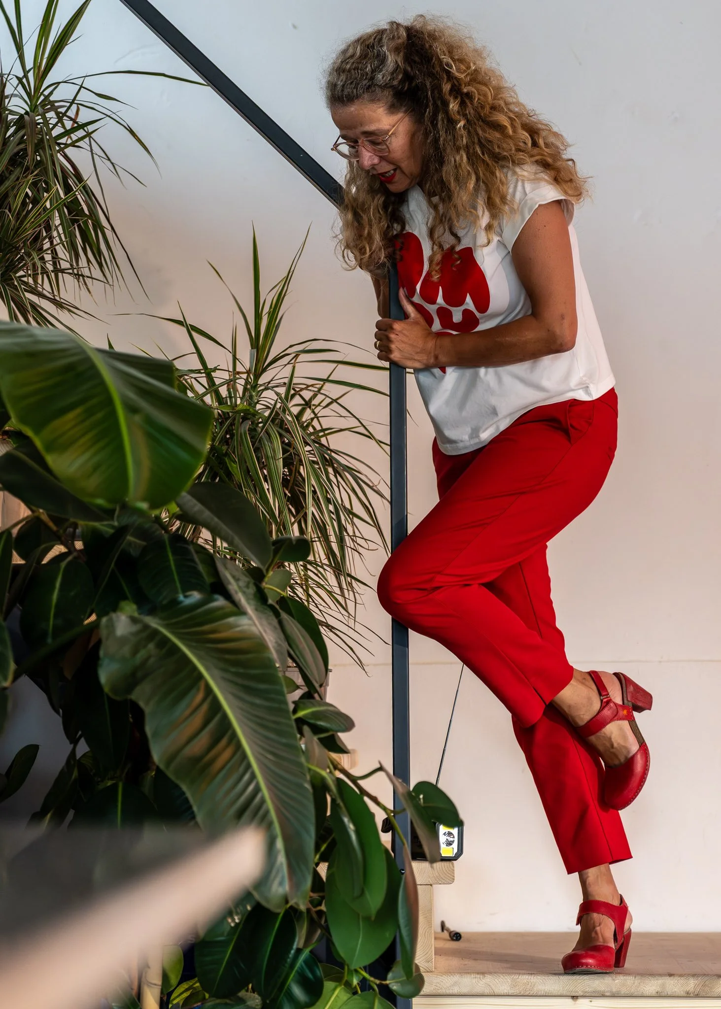 Een vrouw met gekrulde blonde haar draagt een witte T-shirt met rode ontwerp en rode broek, en staat op haar tenen in een kamer met houten vloer en planten.