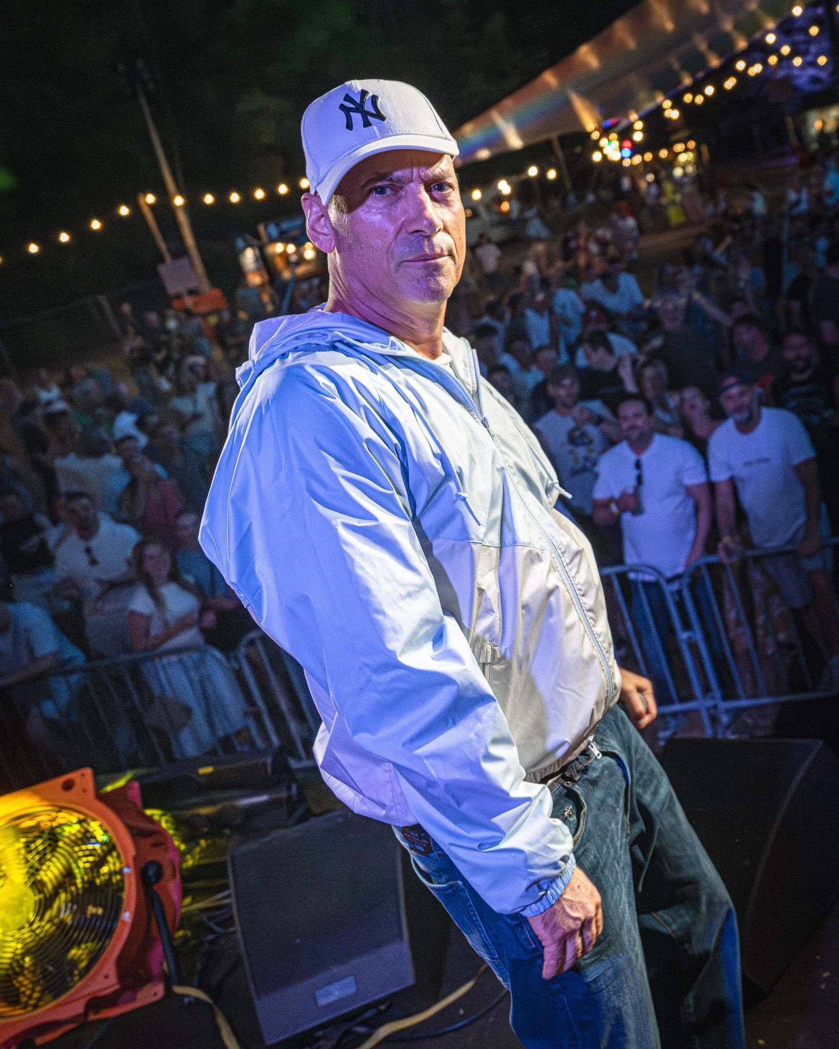 Een man met een wit Yankees-hoed en een witte jas staat in een menigte bij een evenement in de avond. Er zijn mensen op de achtergrond en lichte decoraties. Extince live.