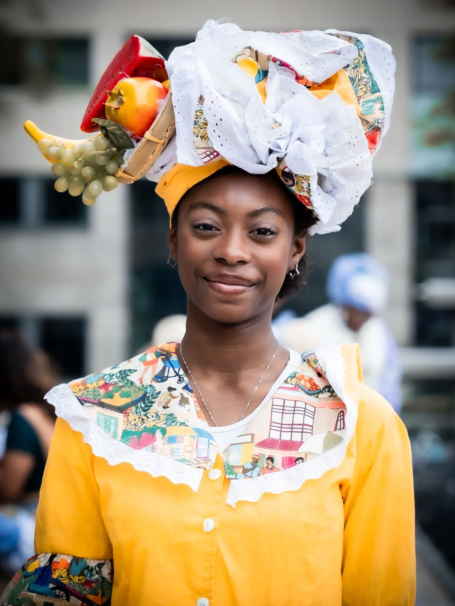 Een vrouw met een hoofdbedekking gemaakt van plastic bakken met speelgoed en fruit, gekleed in een gele outfit met kleurrijke patronen op de mouwen.