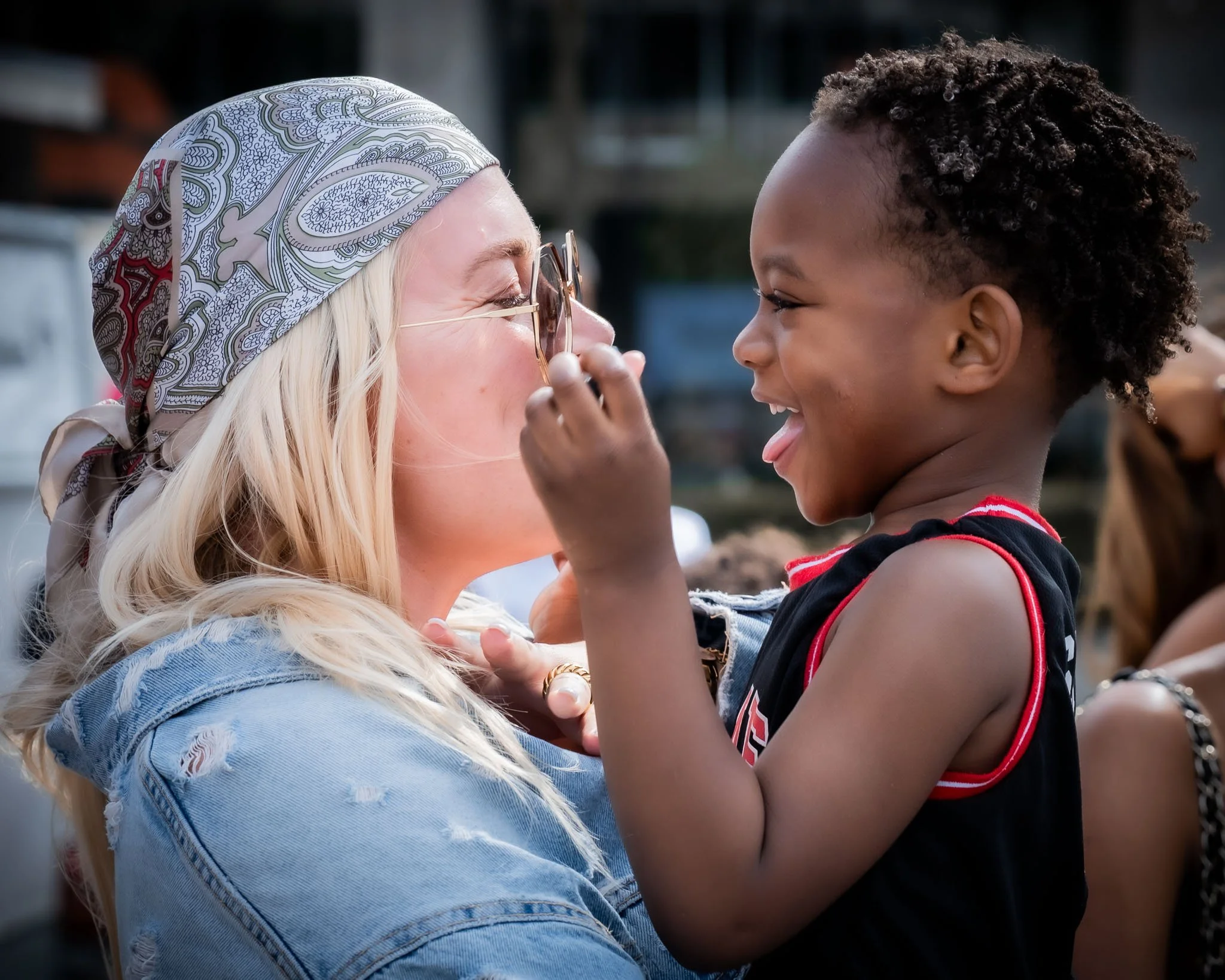 Een vrouw met een geblokte bandana en sieraden die lachend naar een kind kijkt, dat haar vastklampt en haar op de neus tikt.
