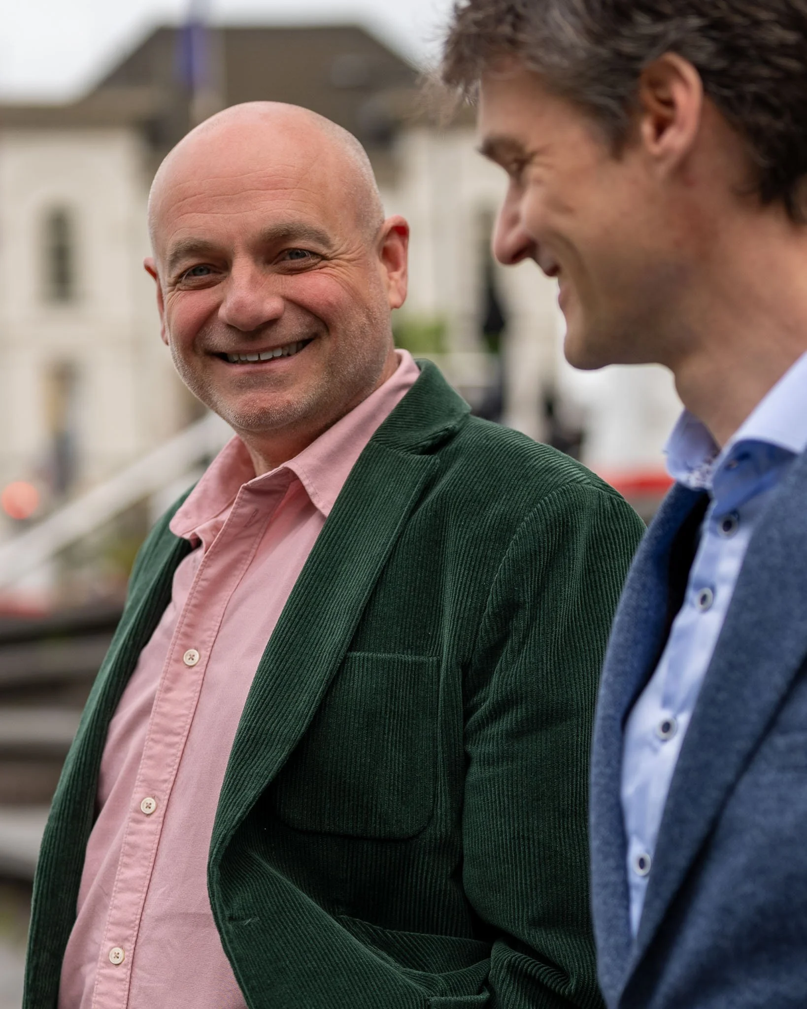 Twee mannen in gesprek, een glimlachend en een lachend, buiten met gebouwen op de achtergrond.