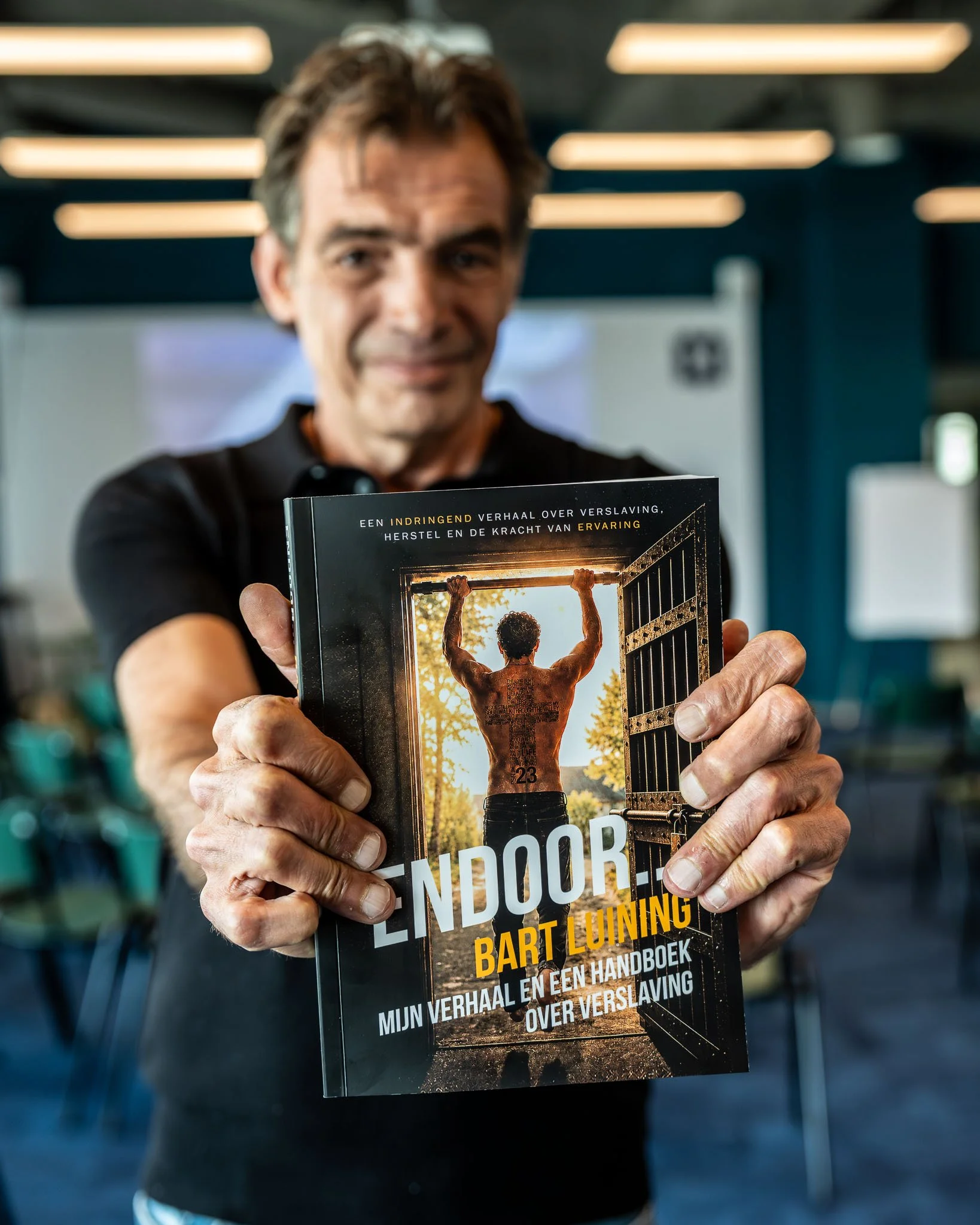 Man houdt een boek voor zich dat de cover toont met een afbeelding van een man die een hek omhoog tilt, onder een boom, tijdens zonsopgang of zonsondergang.