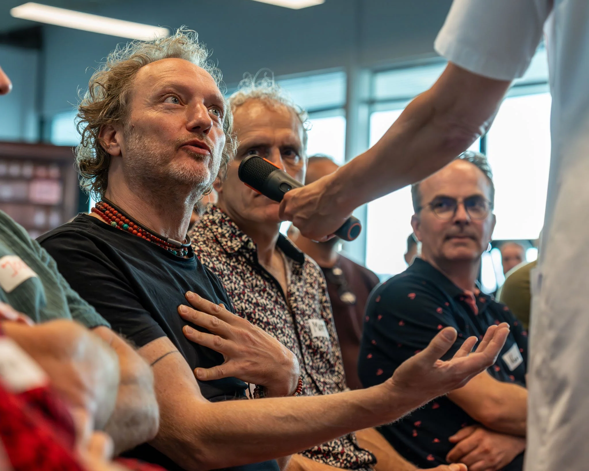 Mensen zitten in een zaal, met één persoon die een microfoon vasthoudt en vragen stelt. Een vrouw met grijs haar en sieraden kijkt aandachtig omhoog.
