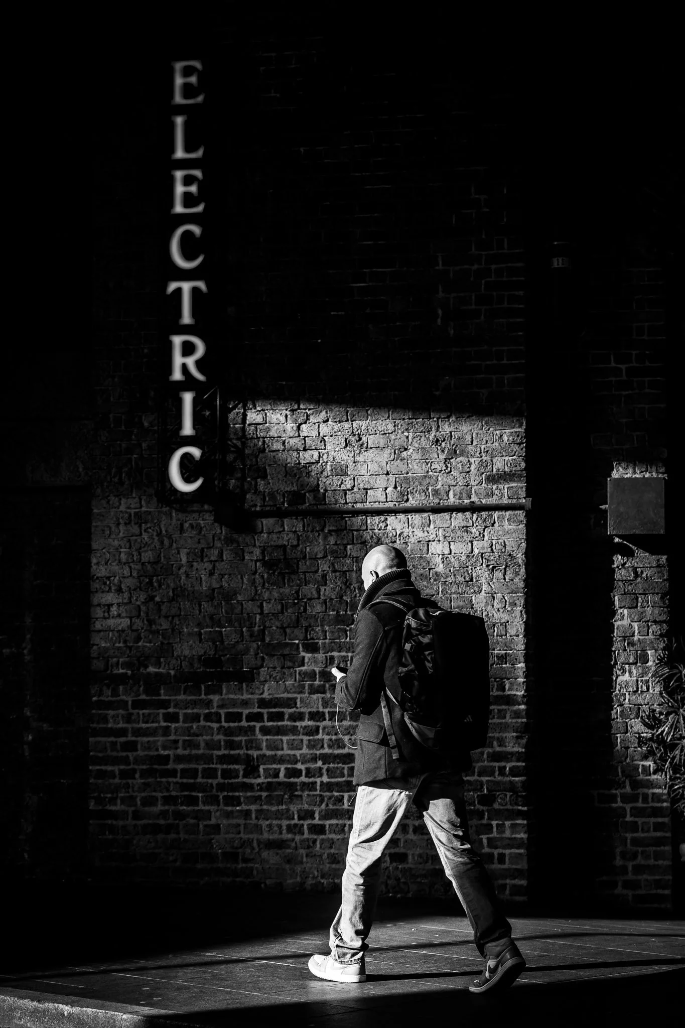 Een man met een rugzak die op een trottoir loopt bij een bakstenen muur met een verlichte verticale tekst 'ELECTRIC'.