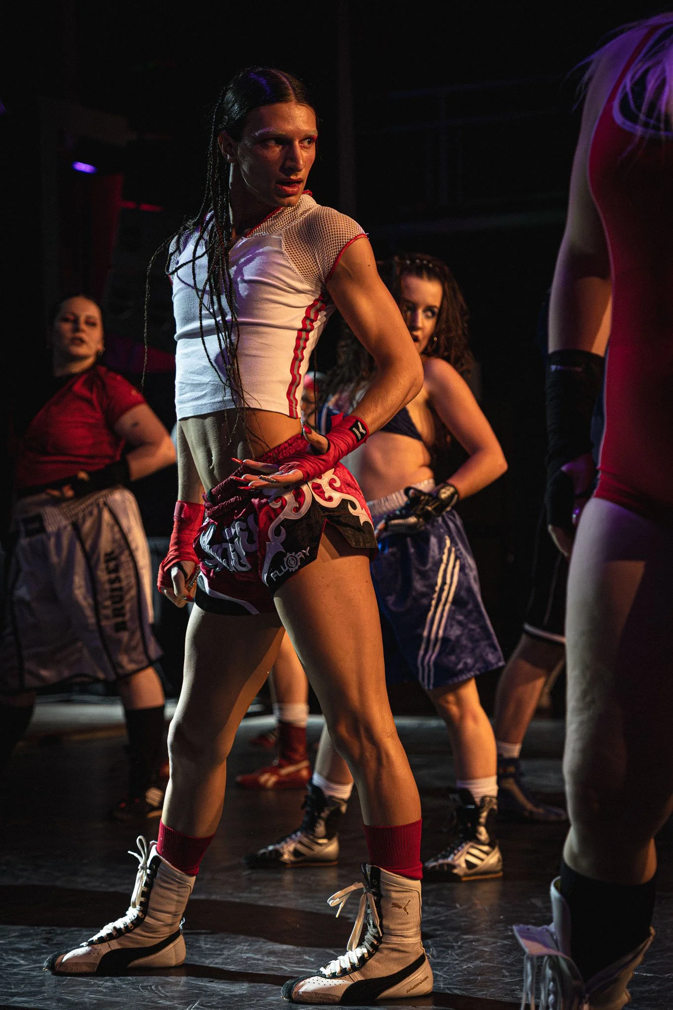 Vrouw in sportoutfit met rollerhockeyschaatsen en dreadlocks, omringd door andere vrouwen, op een donkere podium- of dansvloer.
