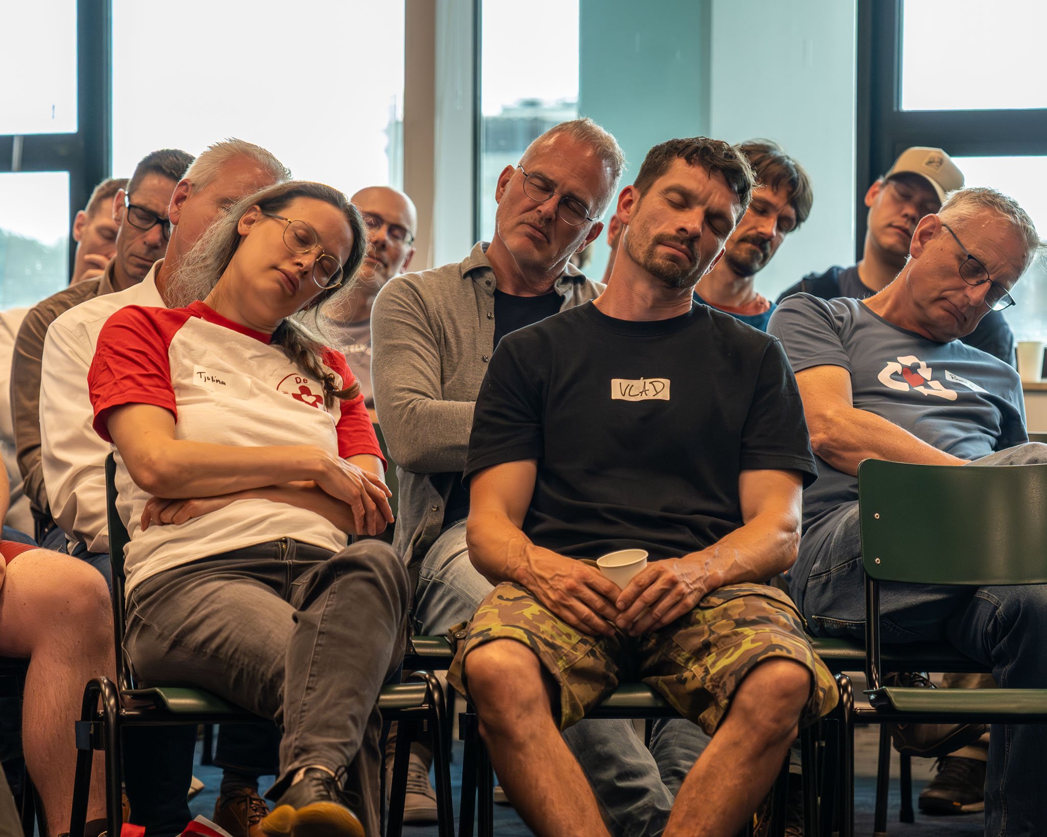 Mensen slapen tijdens een presentatie of conferentie, sommige met hoofden neergeslagen, in een kamer met grote ramen.
