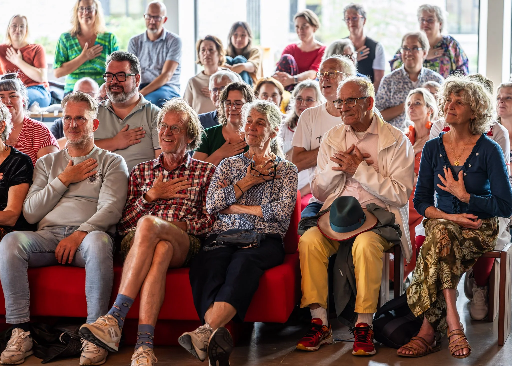 Een groep mensen zit en staat in een zaal, ze lijken te luisteren en te zingen met hun handen op hun hart.