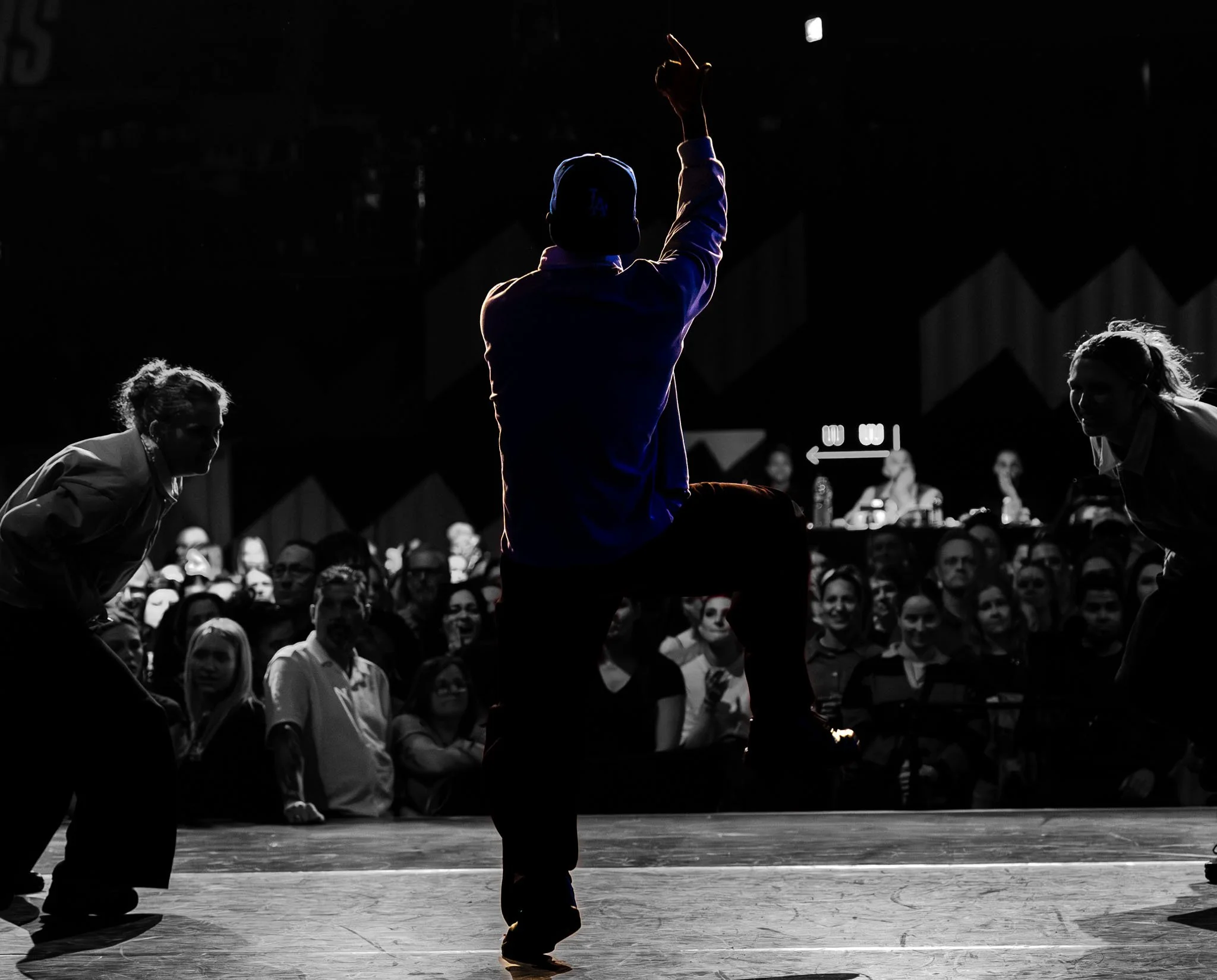 Een silhouet van een persoon met een pet die op een podium danst, met een publiek op de achtergrond.
