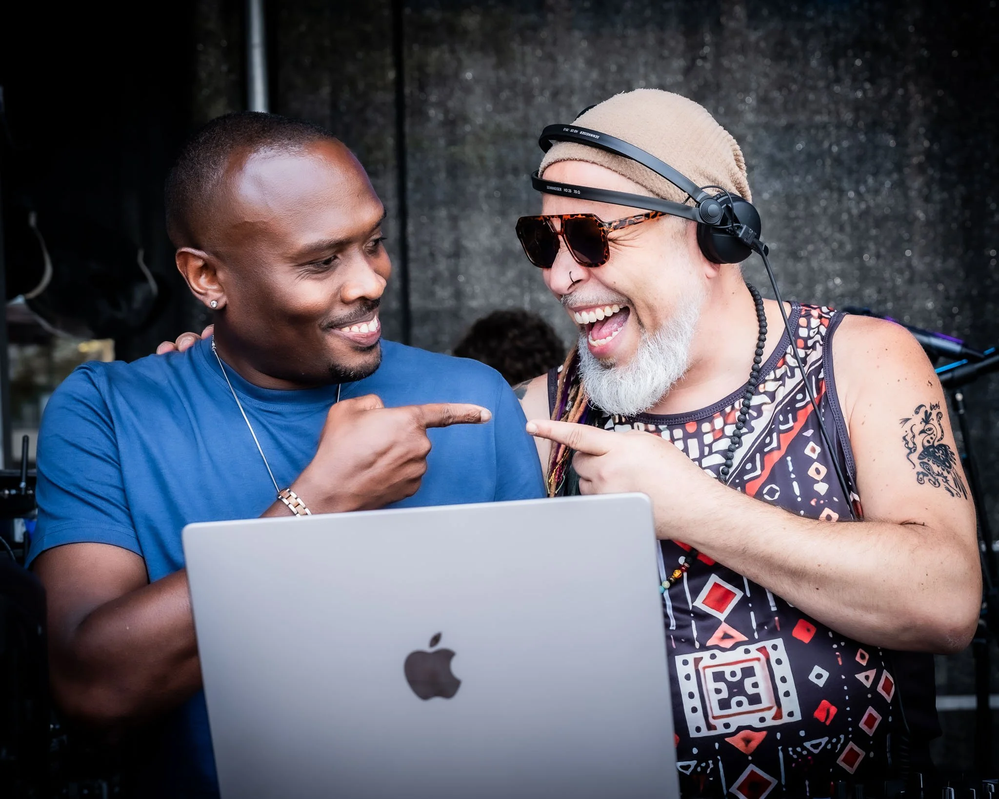 Twee mannen lachen en praten, één wijst naar een laptop, de ander draagt koptelefoon, zonnebril en een beanie, op een donkere achtergrond.