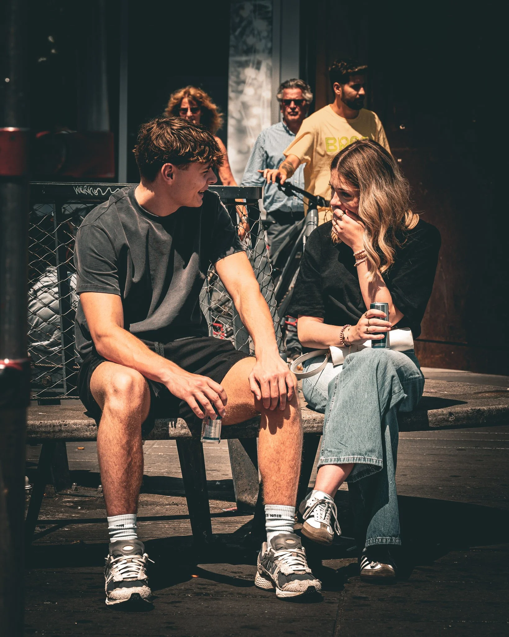 Een jonge man en vrouw zitten op een bankje en lachen samen. Achter hen staan drie mensen en een fiets.
