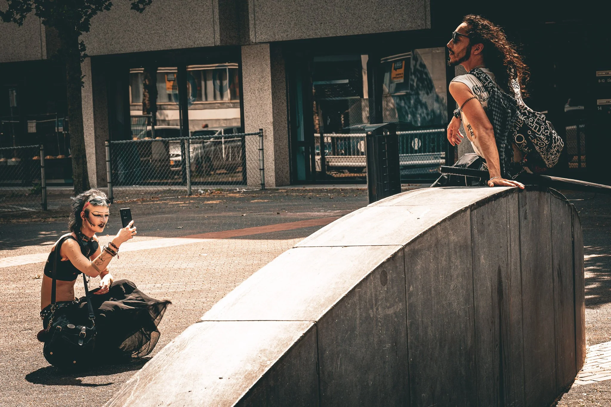 Een man met lang haar en een vrouw met korte haar die foto's maken in een straat met een gebouw en een hek. De man leunt op een rand en draagt een rugzak, de vrouw zit op de grond met een camera en lijkt een selfie te maken.