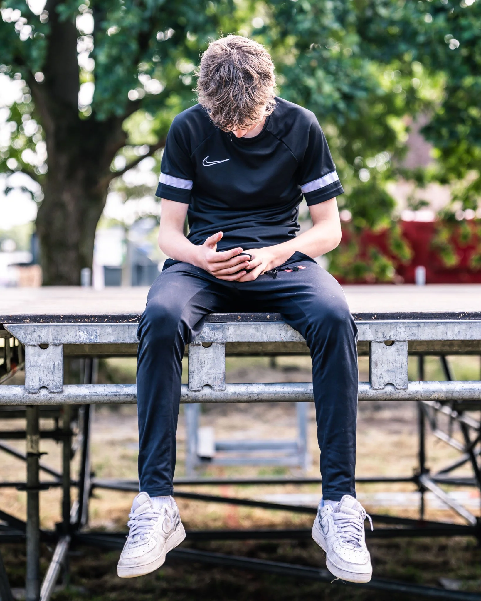 Jongeman met lang haar zit op een bankje onder een boom, kijkt naar beneden, draagt een zwarte Nike sportoutfit en witte sneakers.
