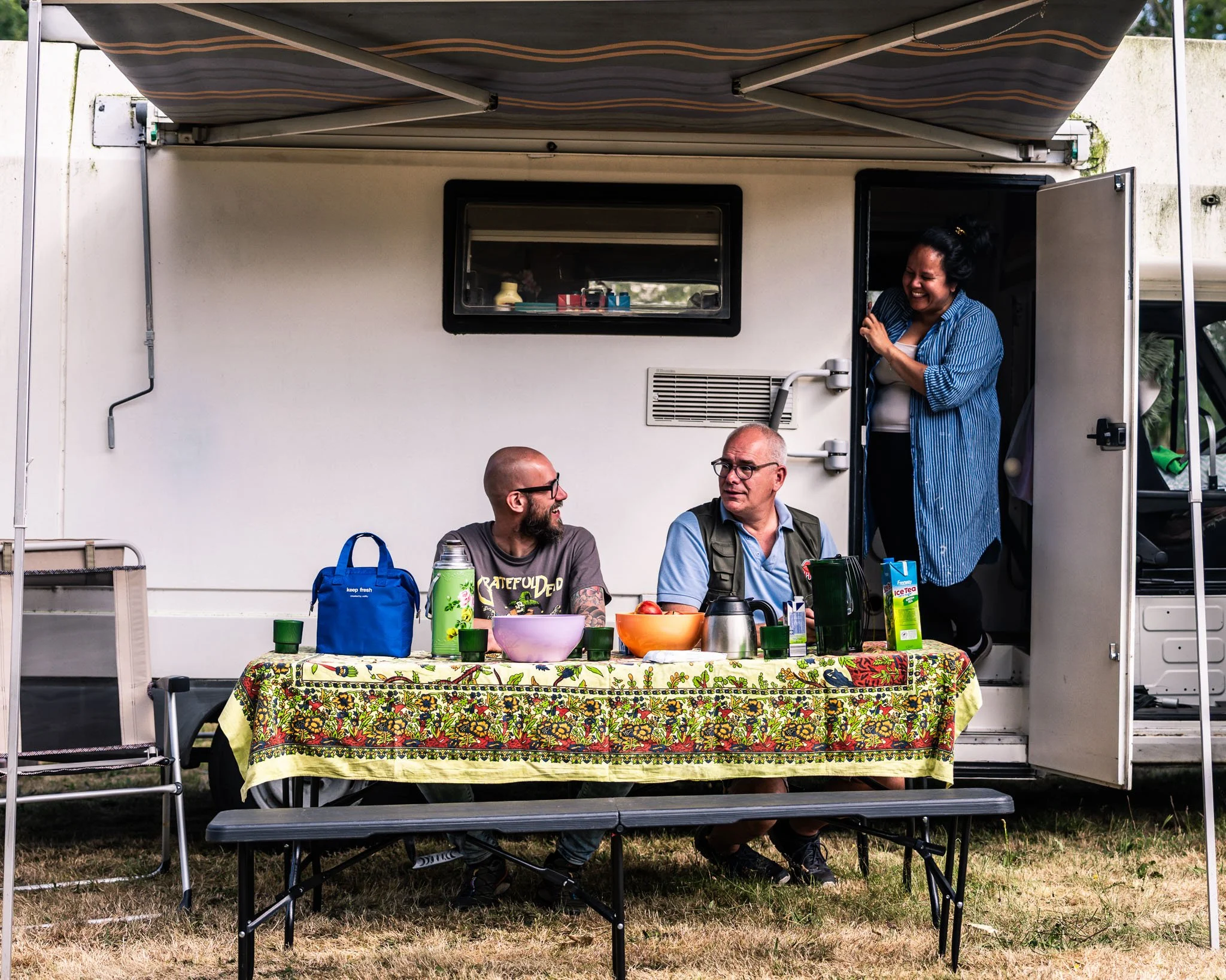 Liefhebbers zitten aan een tafel met een kleurrijk tafelkleed, drankjes en snacks in een camper of caravan tijdens een buitenbijeenkomst, onder een parasol.
