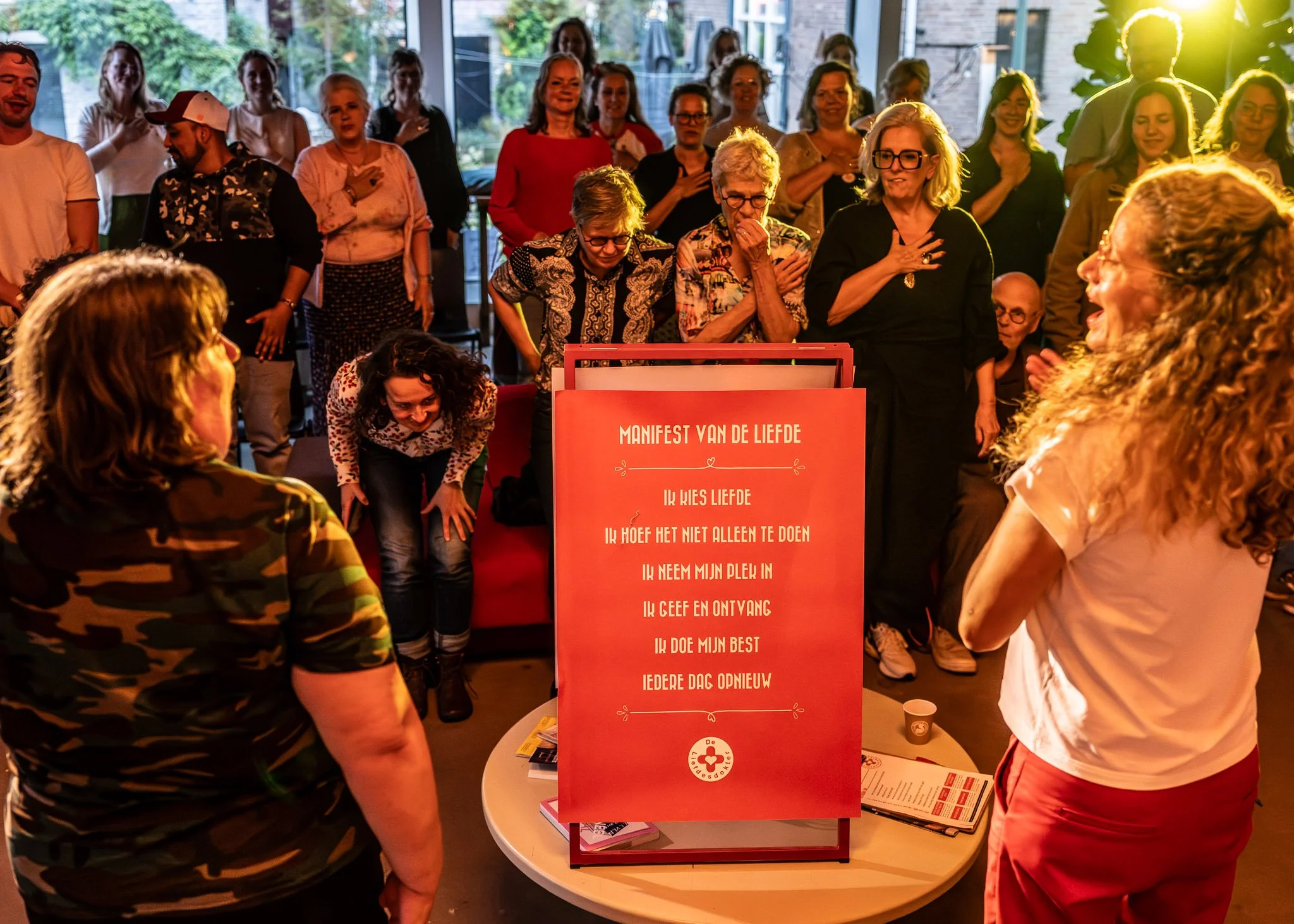 Een groep mensen staat bijeen in een gezellige binnenruimte met grote ramen en groen buitenzicht, terwijl ze naar een rode display met witte tekst kijken en interactie hebben.