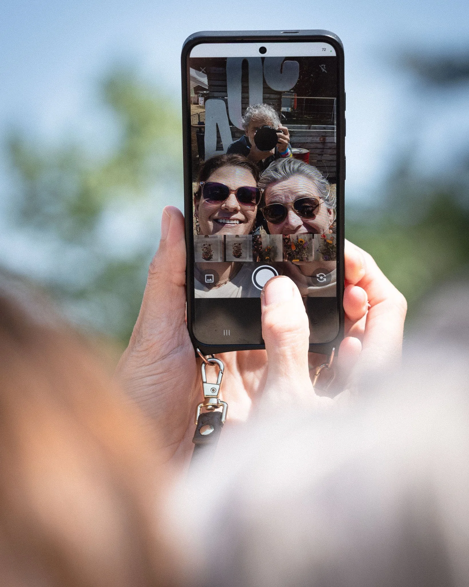 Close-up van een hand die een smartphone vasthoudt en een selfie maakt met twee glimlachende vrouwen met zonnebrillen.