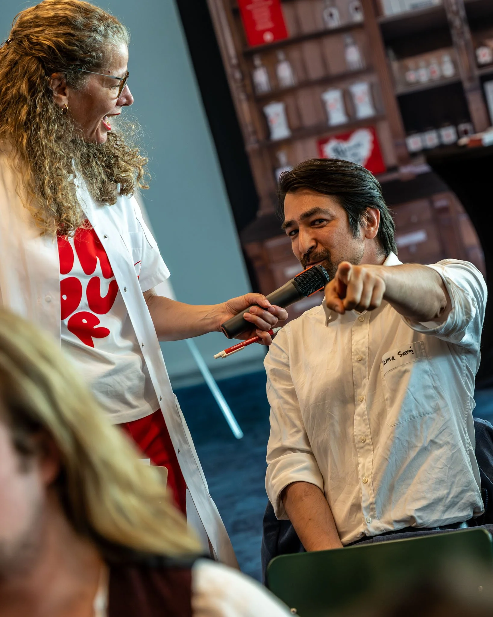 Een vrouw met krullend haar en brillen, lachend en pratend met een man die een microfoon vasthoudt en naar haar wijst, terwijl hij in een witte blouse zit.