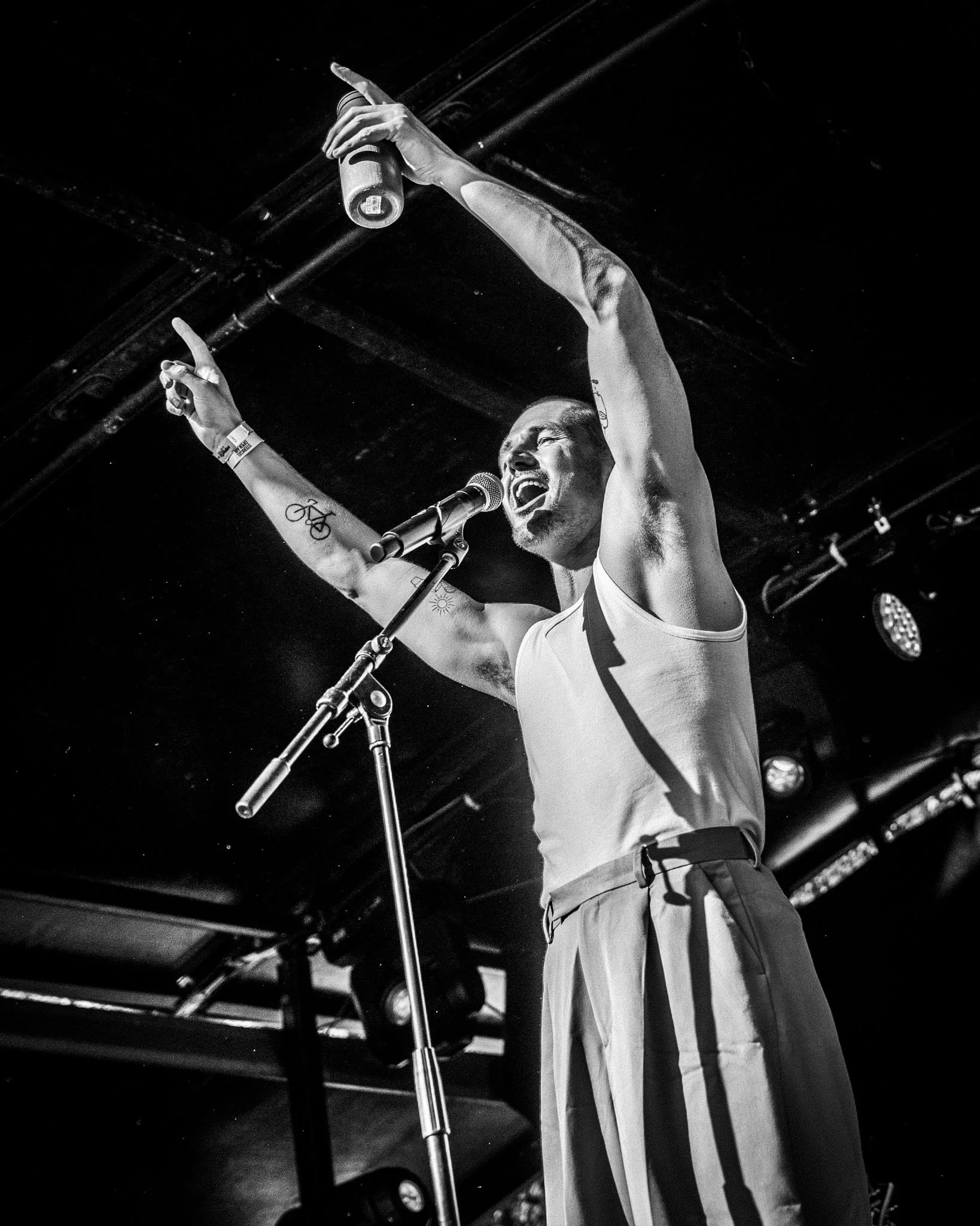 een man die zingt in een microfoon met een arm omhoog en een gebaar met zijn hand, in een donkere ruimte met muziekapparatuur op de achtergrond