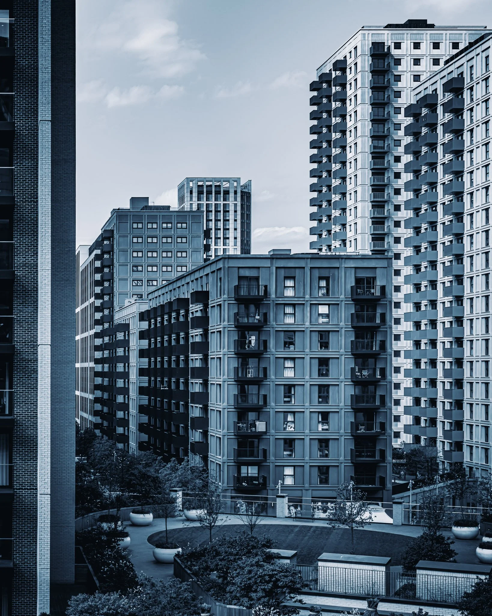 Hoge moderne appartementencomplexen met balkons, een tuin met planten en grote witte vazen, onder een bewolkte hemel.