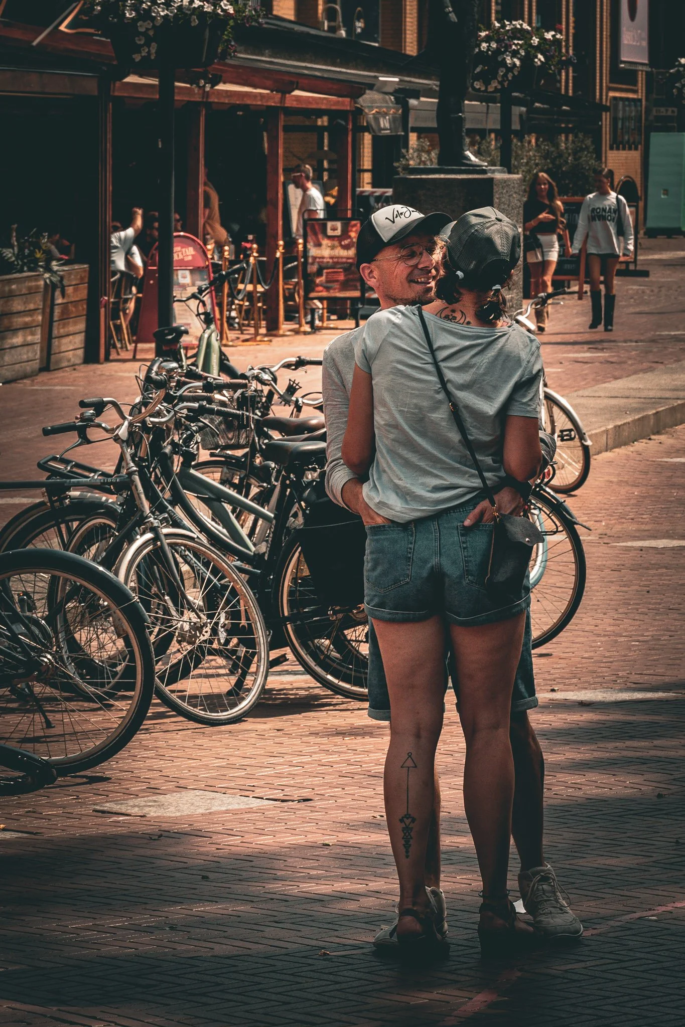 Twee mensen omhelzen elkaar op een fietsenstalling, met fietsen op de voorgrond en een straat met winkels op de achtergrond.