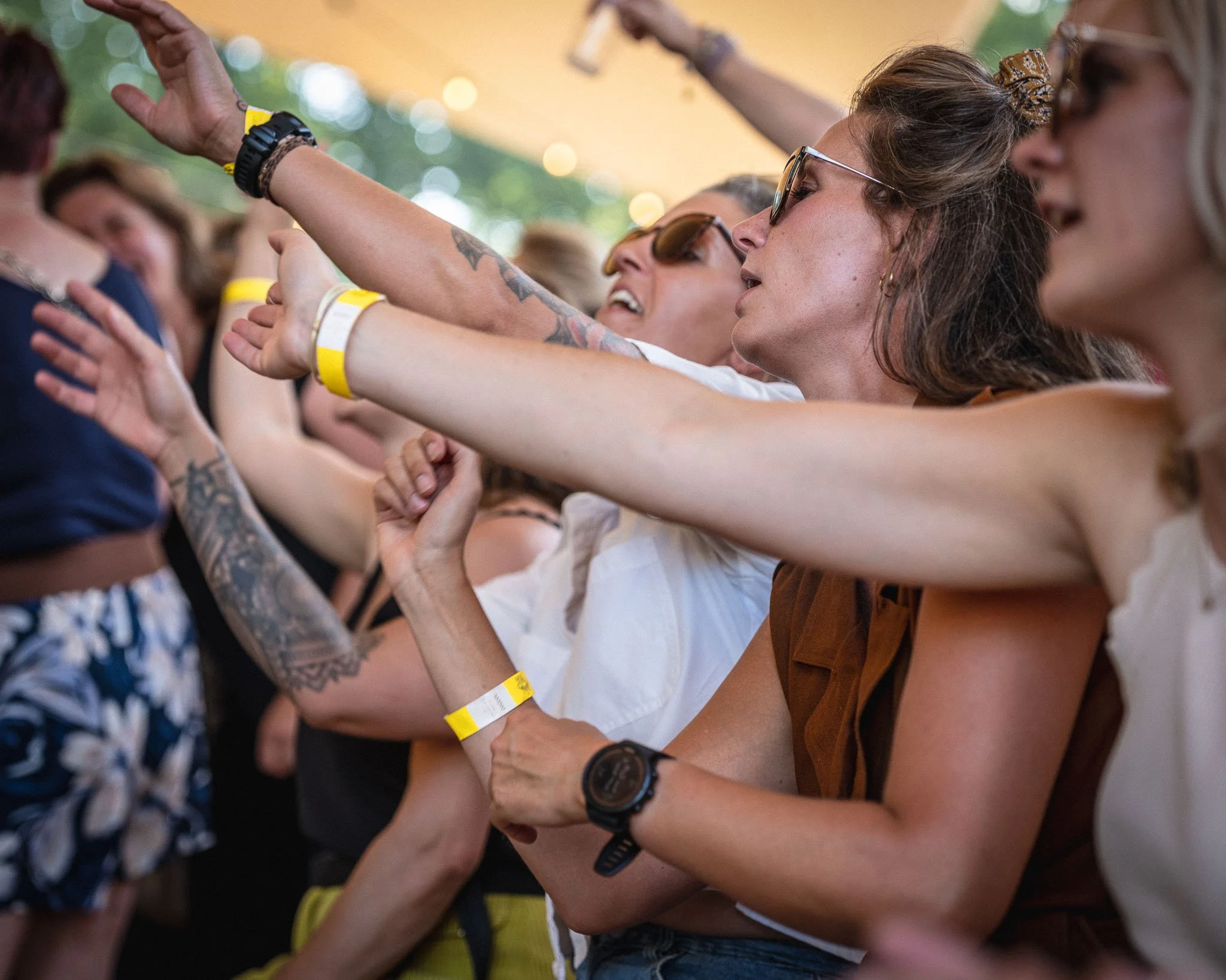 Vrouw met bruin haar en zonnebril die haar arm uitstrekt, omringd door andere mensen met armbanden op een evenement.