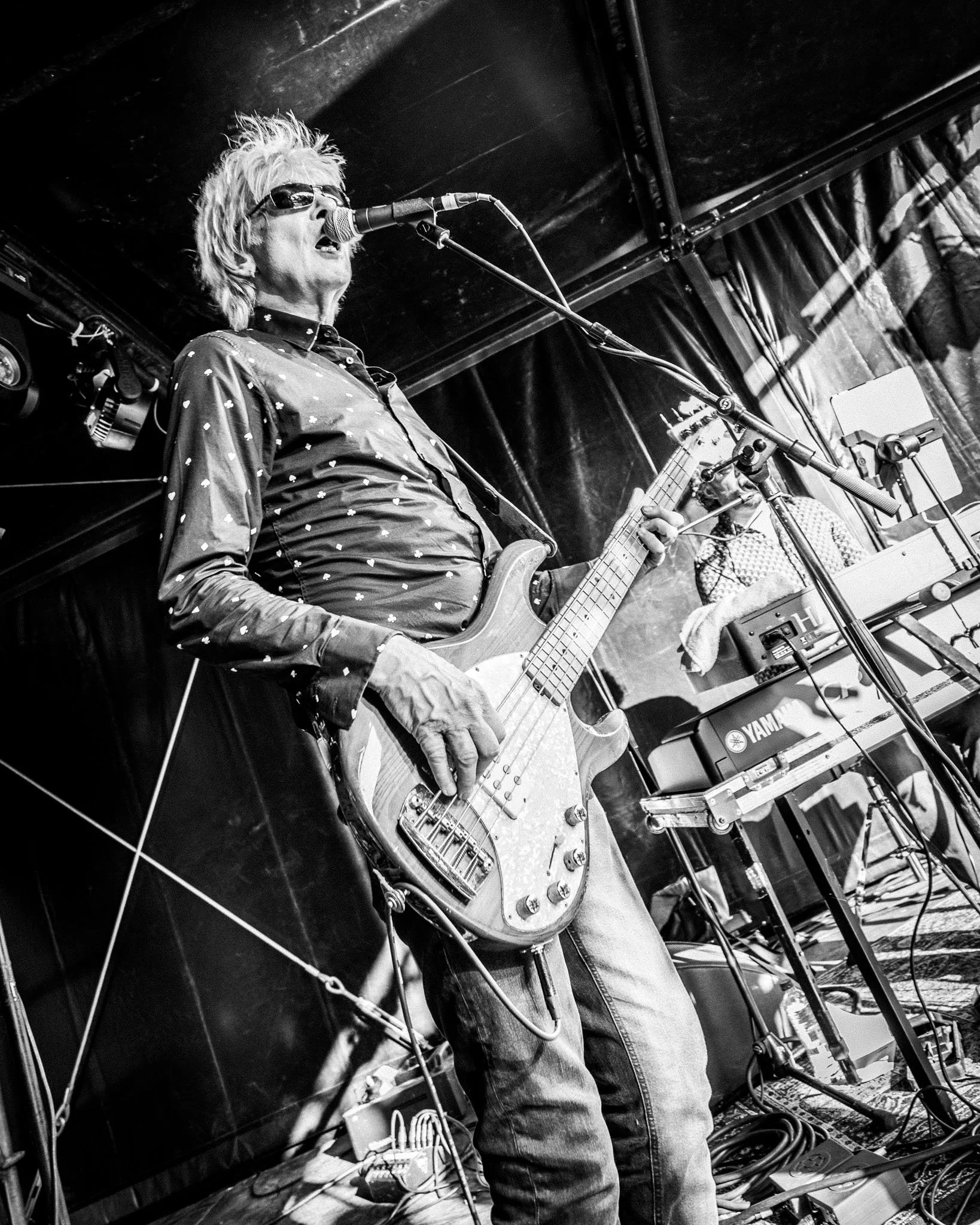 Een man met wit haar en zonnebril speelt elektrische gitaar en zingt in een zwart-wit foto tijdens een optreden, met een toetsenist op de achtergrond, onder een zwarte plafond en tussen audiotechniek en microfoons.