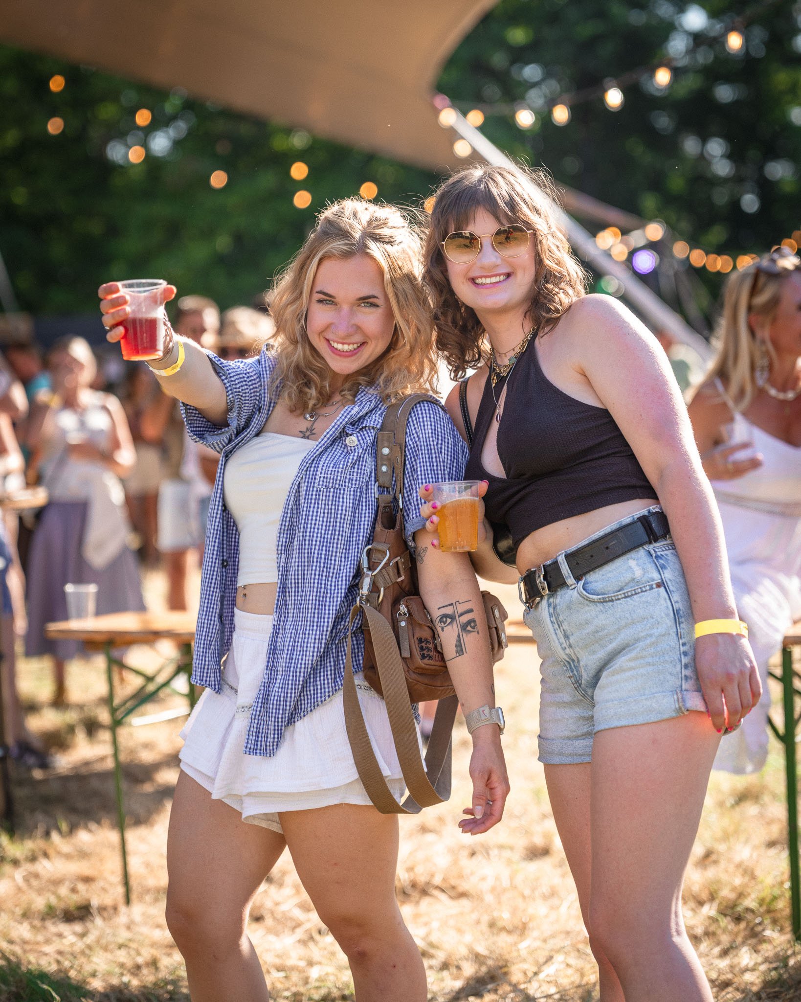 Twee jonge vrouwen genieten op een zomers feest, ze lachen en heffen hun glazen met drankjes omhoog terwijl ze samen poseerden, op de achtergrond zijn andere feestgangers en lichte decoraties te zien.