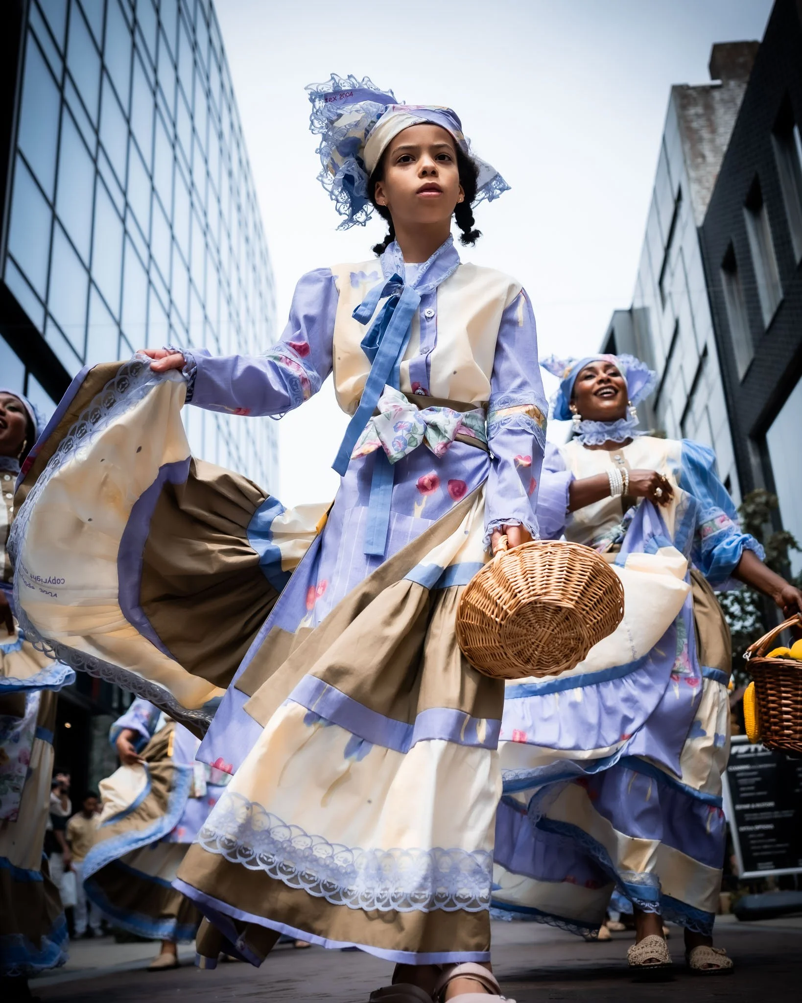 Vrouw in traditionele geklede jurk danst tijdens een straatparade, omringd door andere dansers in vergelijkbare kleding, met moderne gebouwen op de achtergrond.
