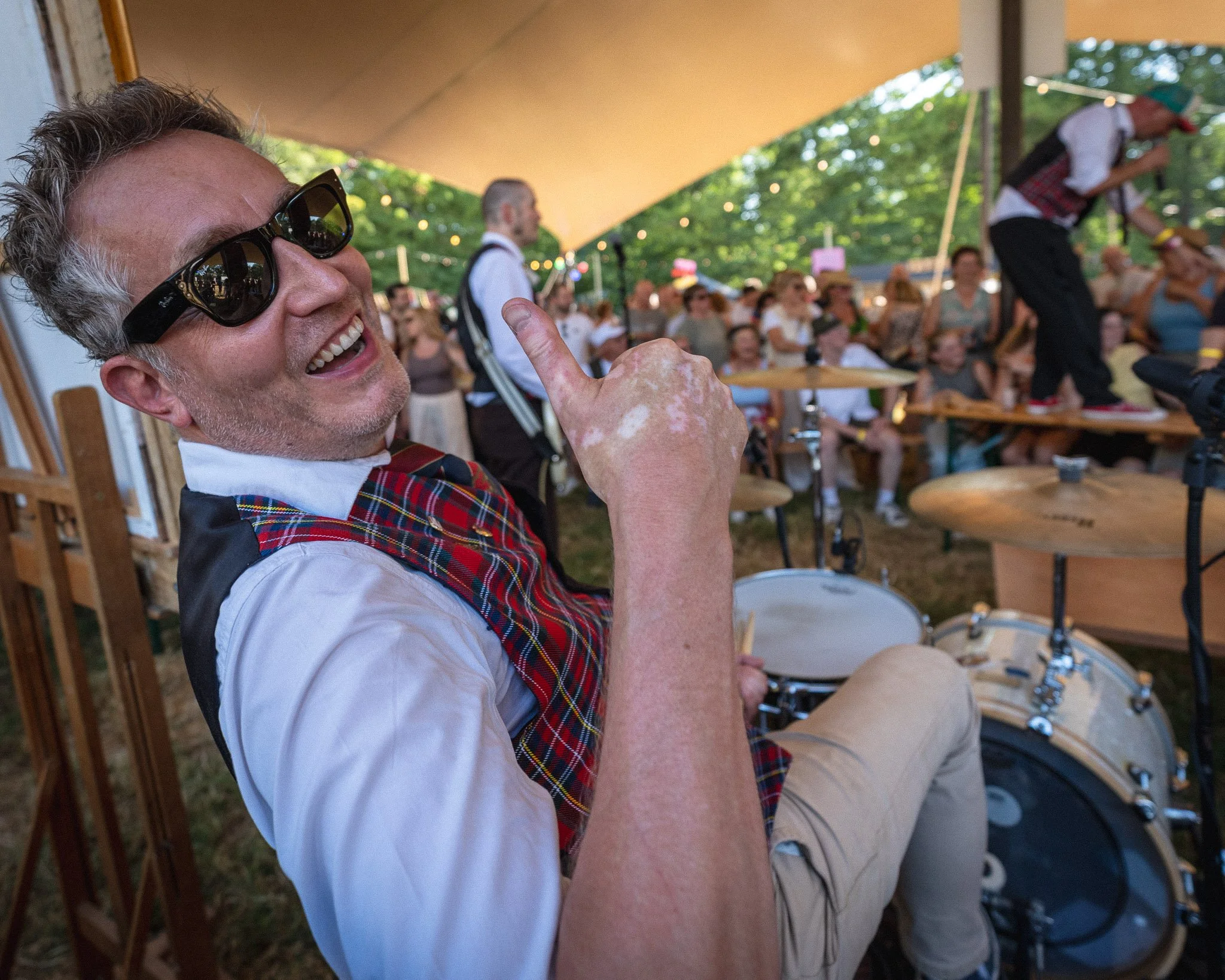 Man met grijs haar en zonnebril speelt op een drumstel onder een grote beige tent, met een menigte mensen op de achtergrond.