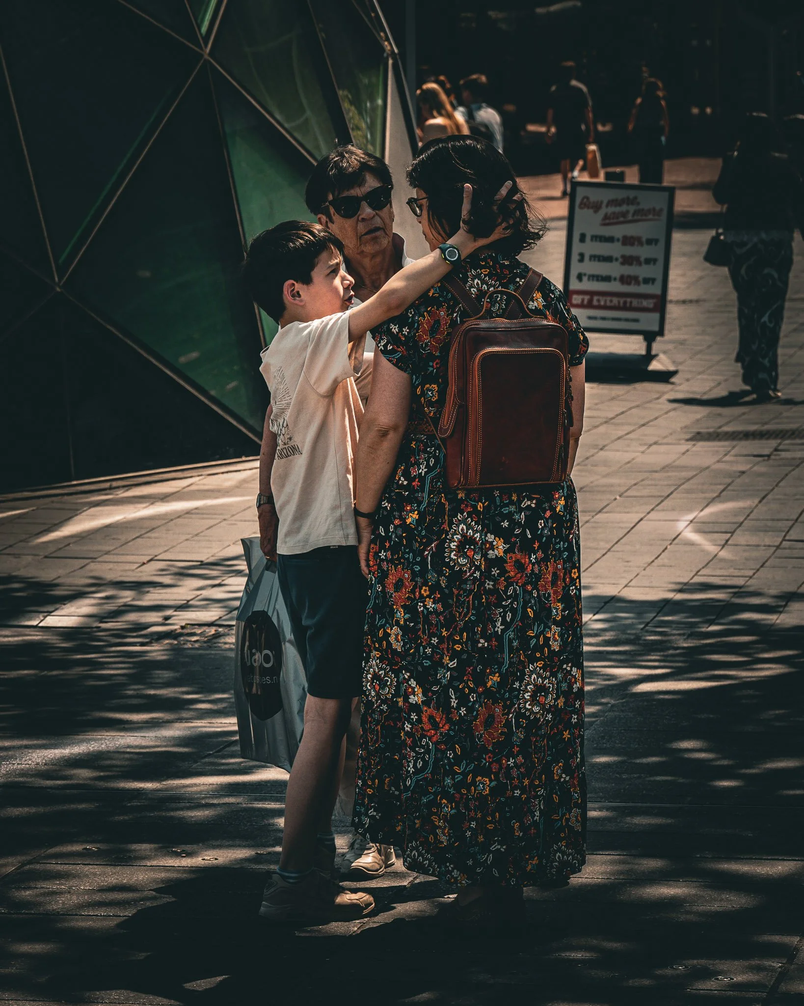 Een kind dat een vrouw omhelst op straat, met een man in de achtergrond. Het lijkt een emotiesessie of een intiem moment tussen hen.