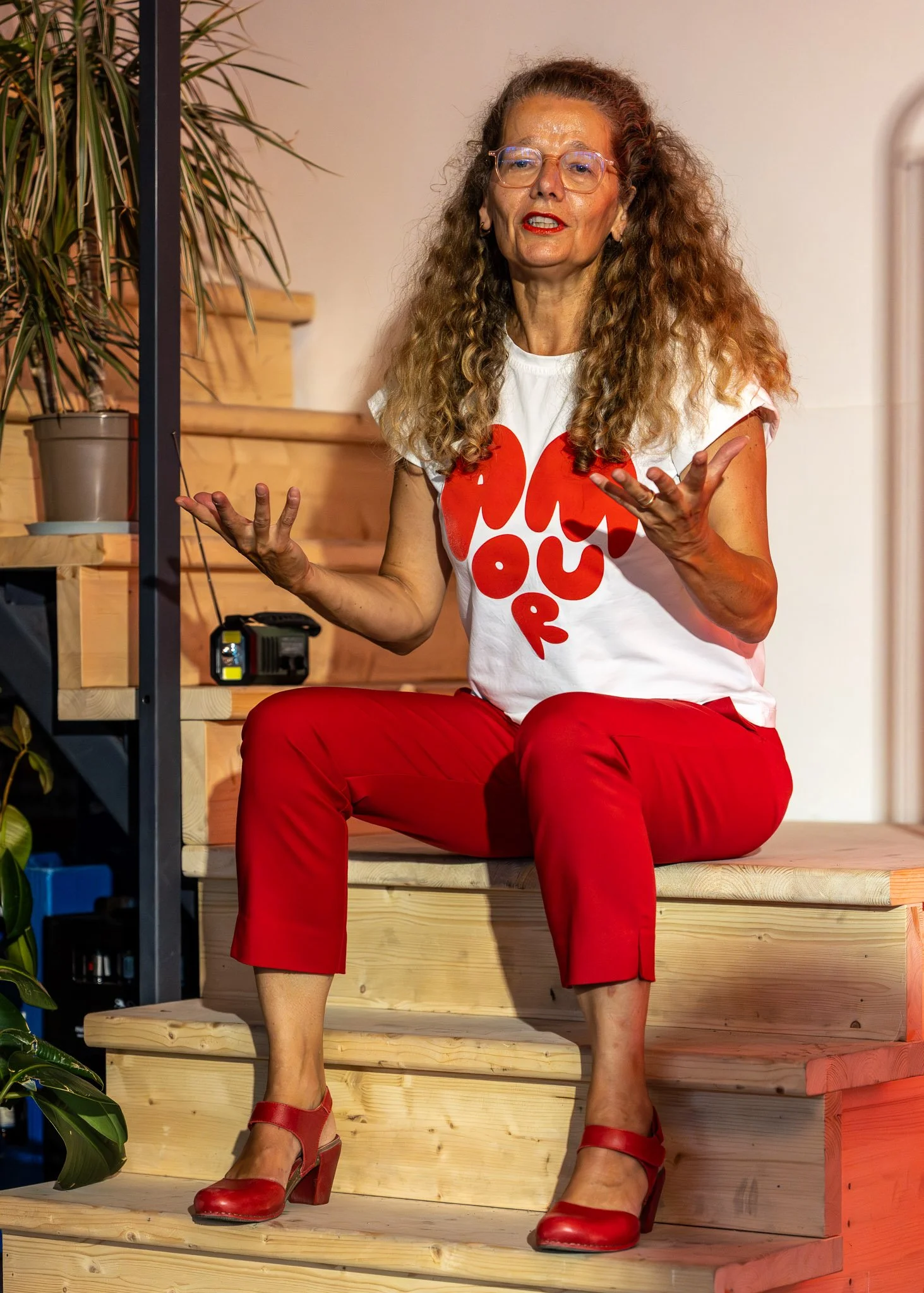 Een vrouw met lang, gekruld haar en glazen, zit op houten trap tachtig met rode schoenen en een witte T-shirt met grote rode tekst, haar handen gebaren terwijl ze spreekt.