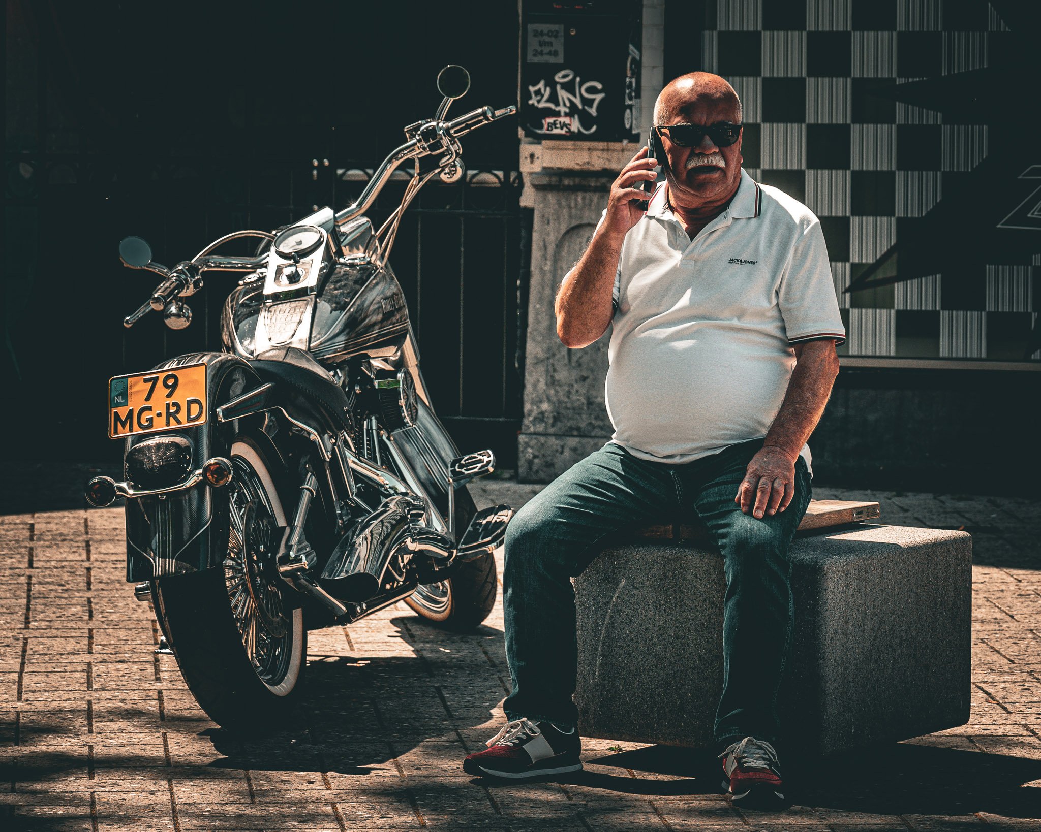 Een oudere man met een witte polo, donkere zonnebril, zittend op een muurtje naast een motorfiets, in een stedelijke omgeving met graffiti en een muur met een geometrisch patroon.