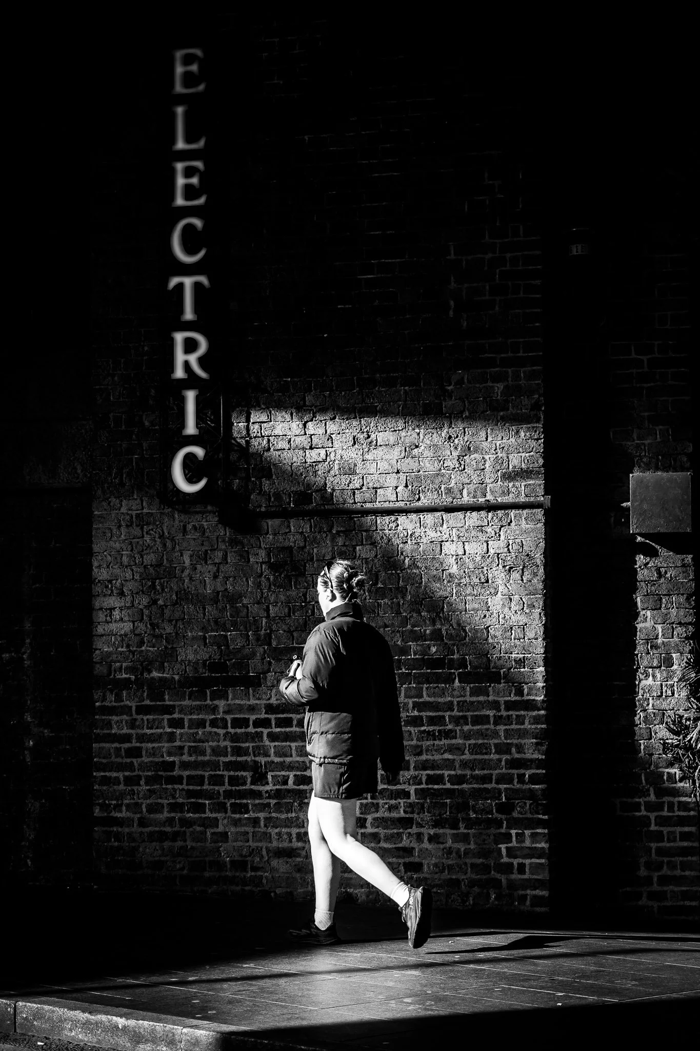 Een persoon die in een zwart-witte foto langs een bakstenen muur loopt, met een lamp die het woord 'ELECTRIC' uitstraalt.