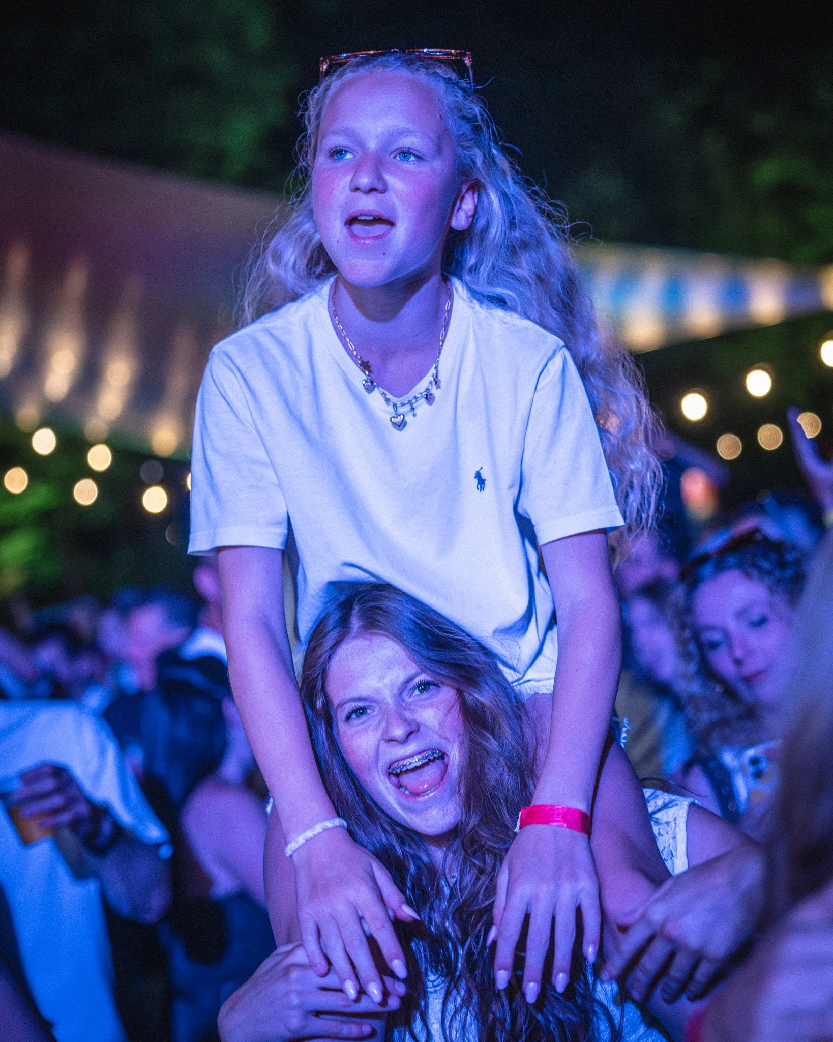 Twee jonge meisjes zijn op een feestje, lachen en hebben plezier. Eén meisje zit op de rug van het andere meisje, dat haar stevig vasthoudt. De achtergrond is donker met kleurrijke lichteffecten en andere mensen die feest vieren.