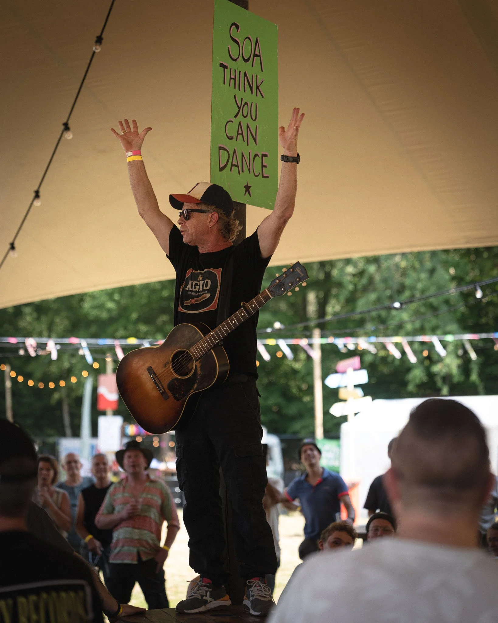 Man met een gitaar staat op een podium onder een tent en houdt een groen teken met de tekst 'SOA THINK YOU CAN DANCE'. Mensen kijken naar hem en er hangen lampjes en vlaggetjes in de achtergrond.